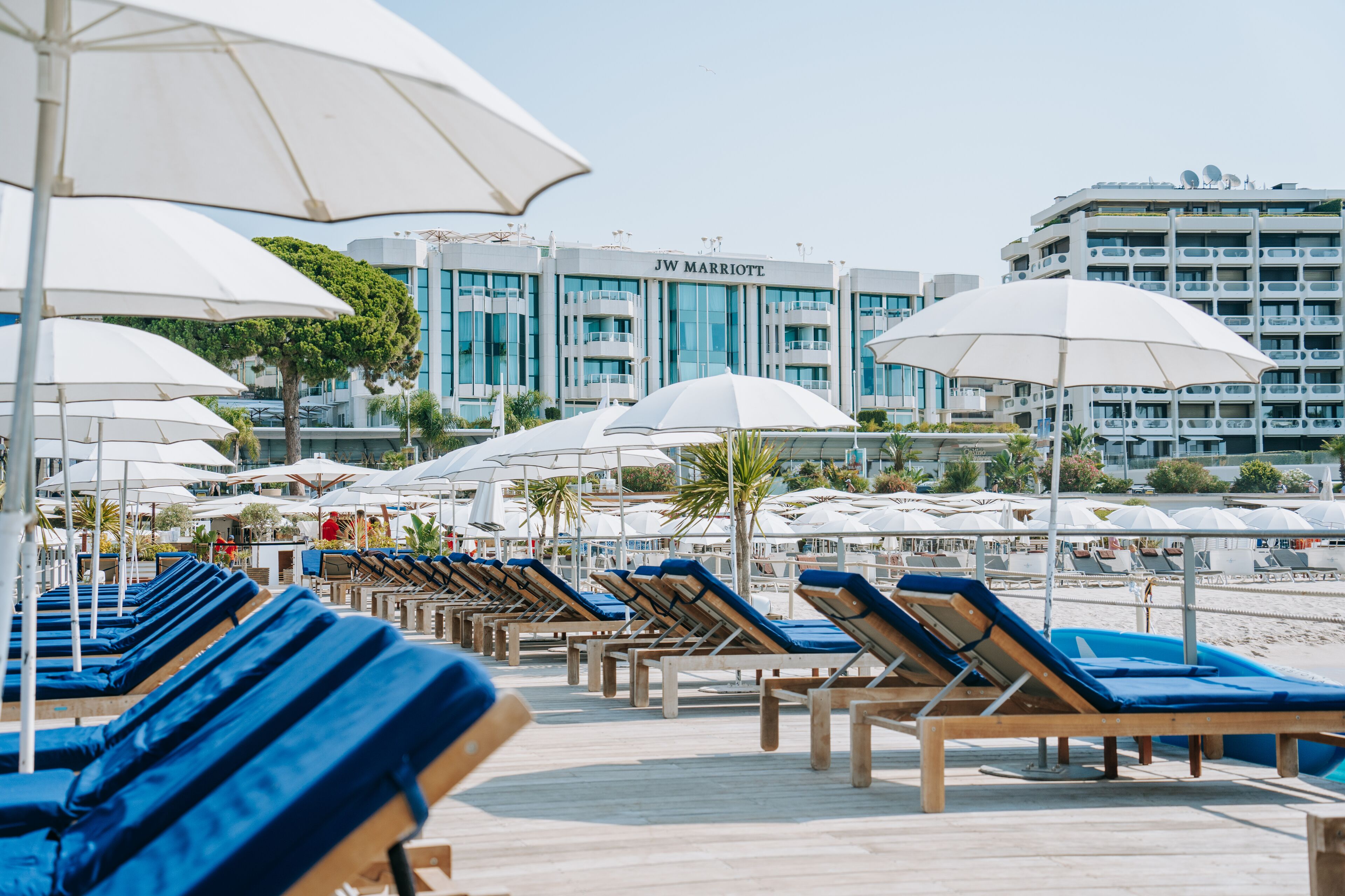 A beachside hotel with rows of sun loungers and umbrellas, a JW Marriott building in the background, and a clear sky.