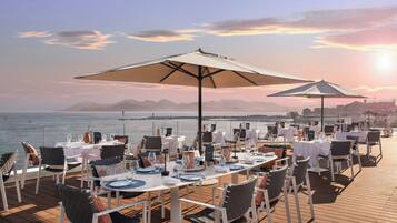 An outdoor dining area with tables and chairs, overlooking a body of water and a cityscape in the distance.