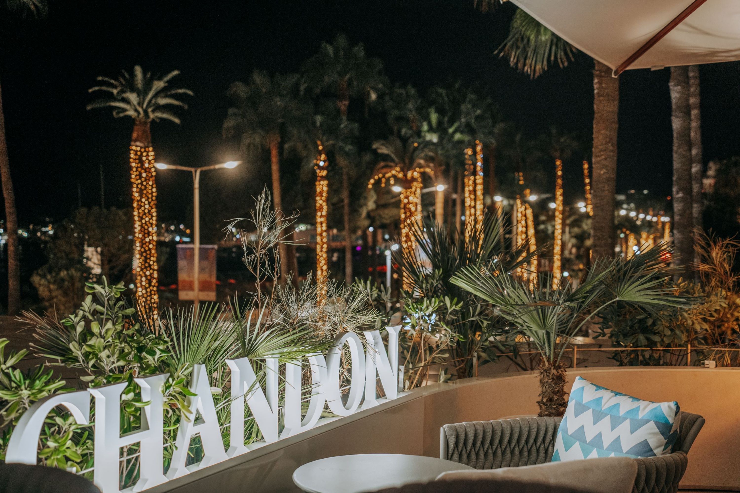 Un área de asientos al aire libre de noche con palmeras, luces decorativas y un letrero que dice 'CHANDON'.