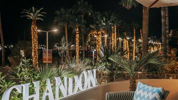 Un área de asientos al aire libre de noche con palmeras, luces decorativas y un letrero que dice 'CHANDON'.