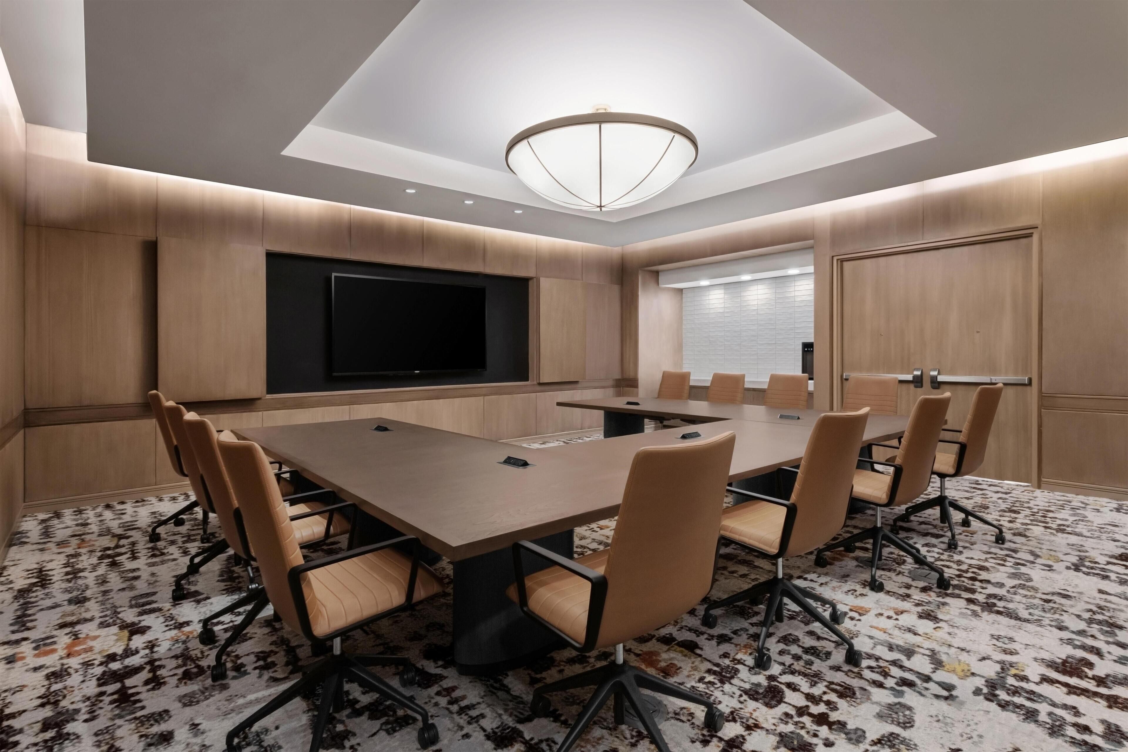 A conference room with a large table, chairs, a flat-screen TV, and a patterned carpet.