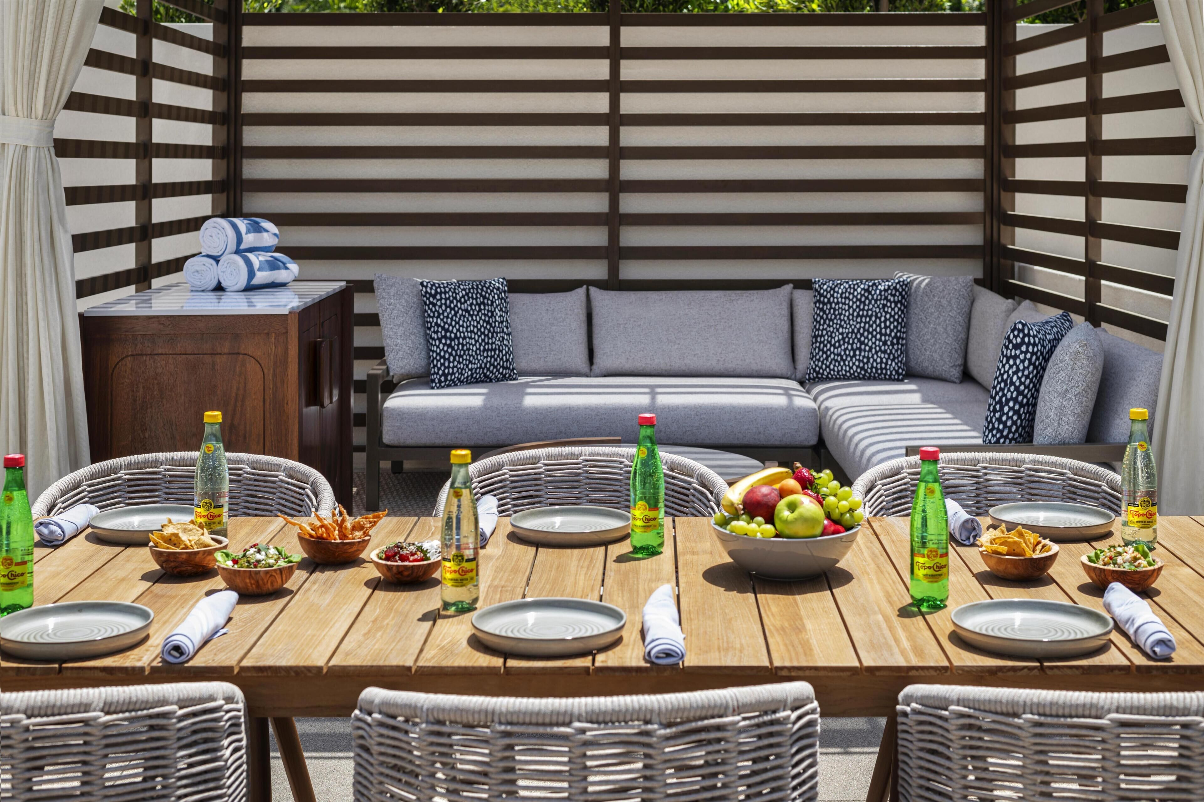 An outdoor dining area with wicker chairs, a wooden table set with plates, bowls, and bottles, and a cushioned seating area with pillows.