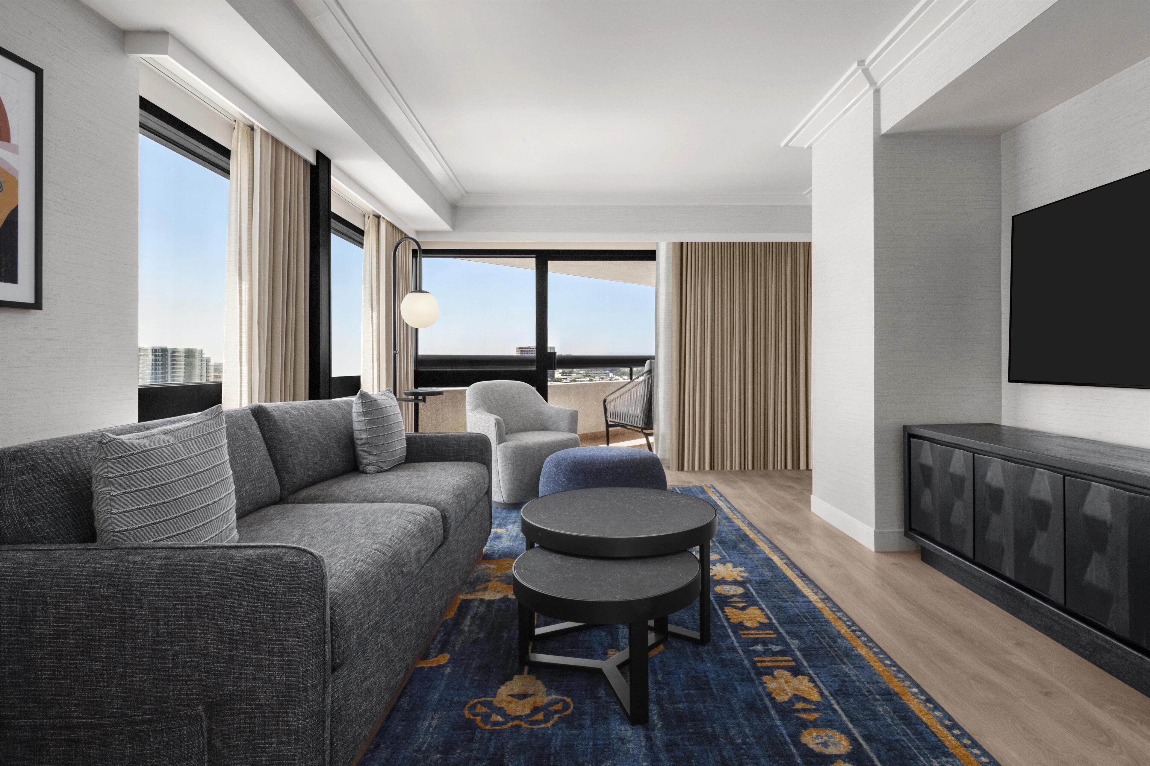 A modern hotel room with a grey sofa, a round table, a TV, and a view of the city.