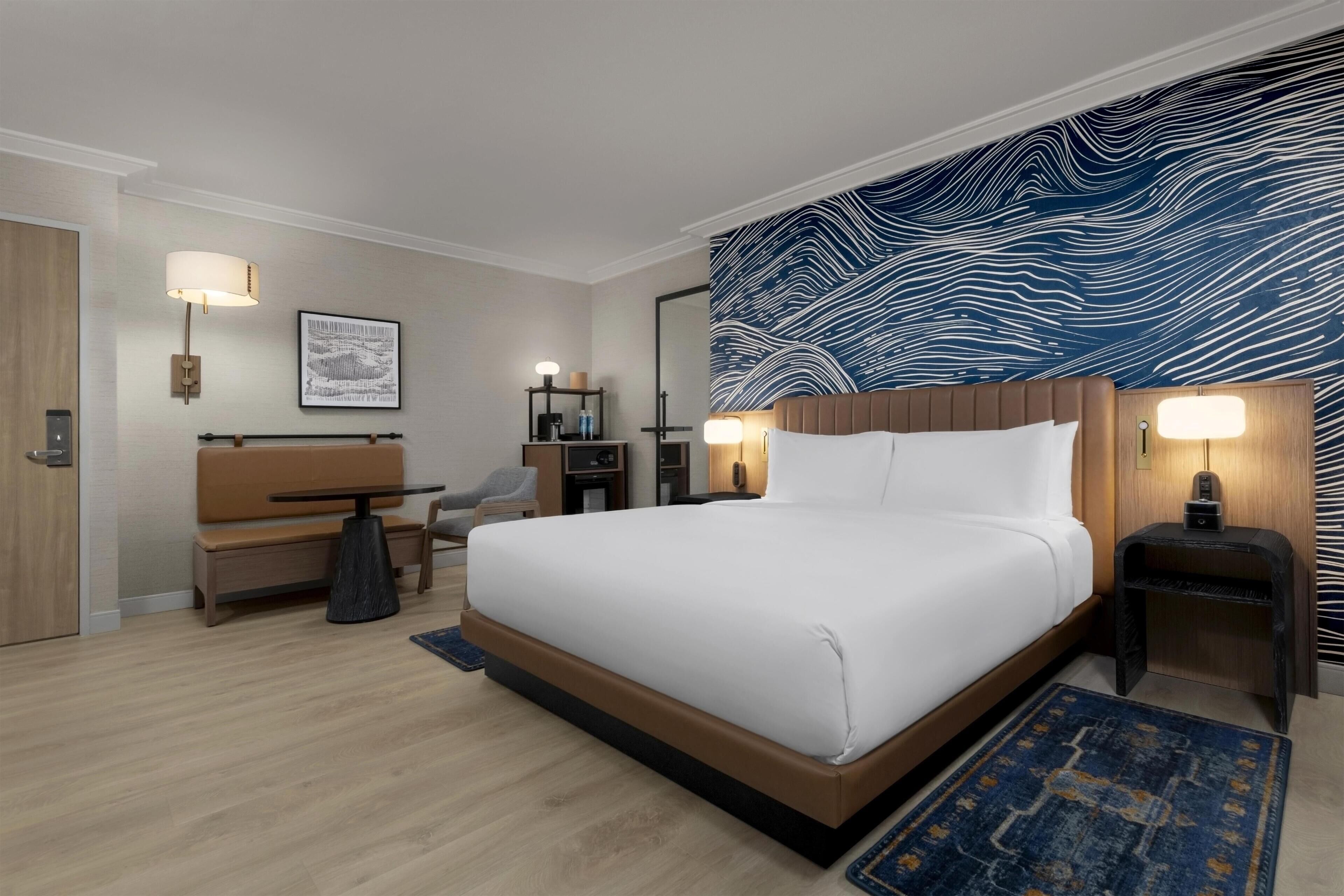 Une chambre d’hôtel moderne avec un grand lit, un bureau, une chaise, une petite table et un mur orné d’un motif ondulé.