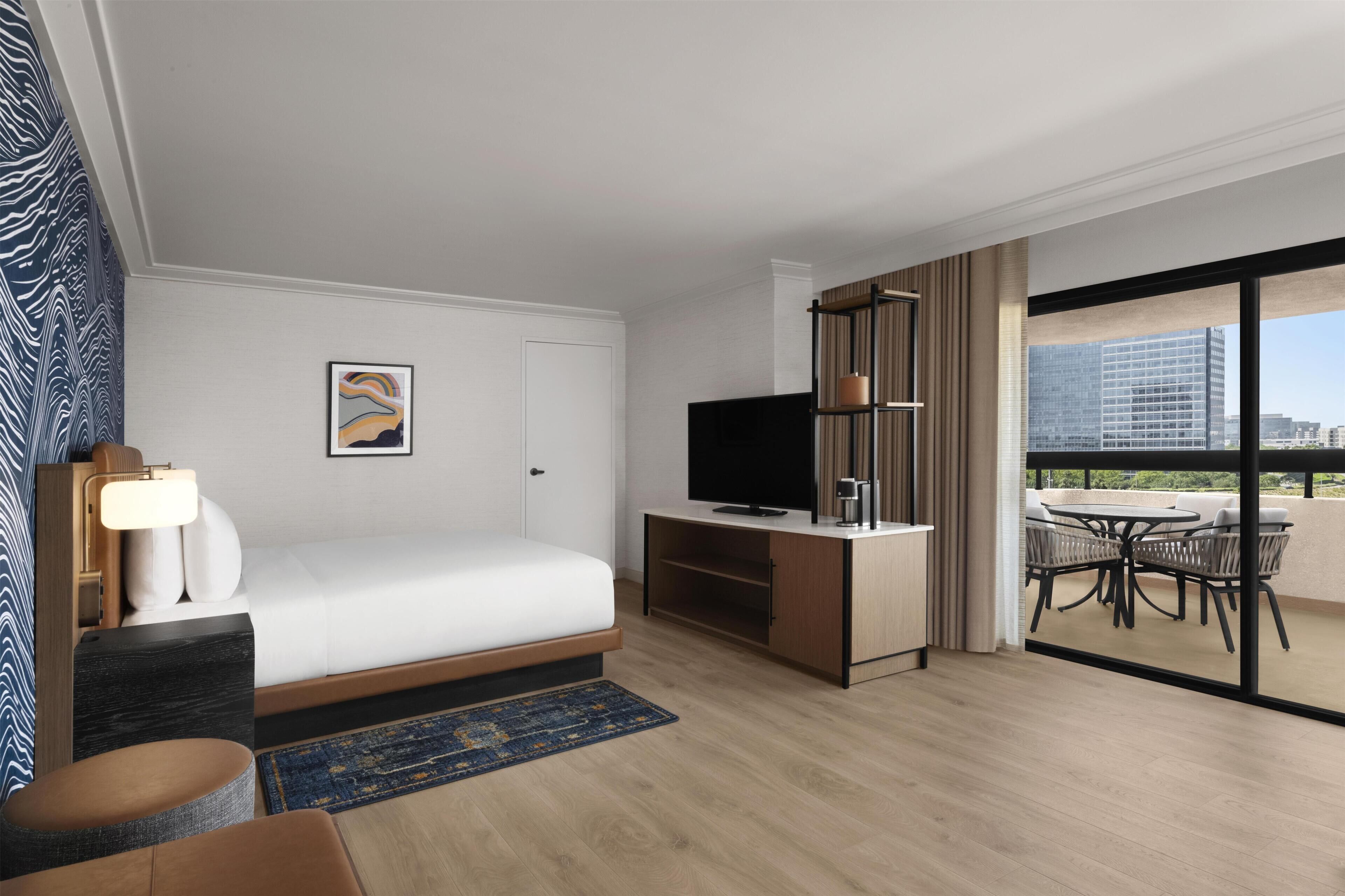 A hotel room with a bed, a desk, a TV, a balcony with outdoor furniture, and a view of the city.