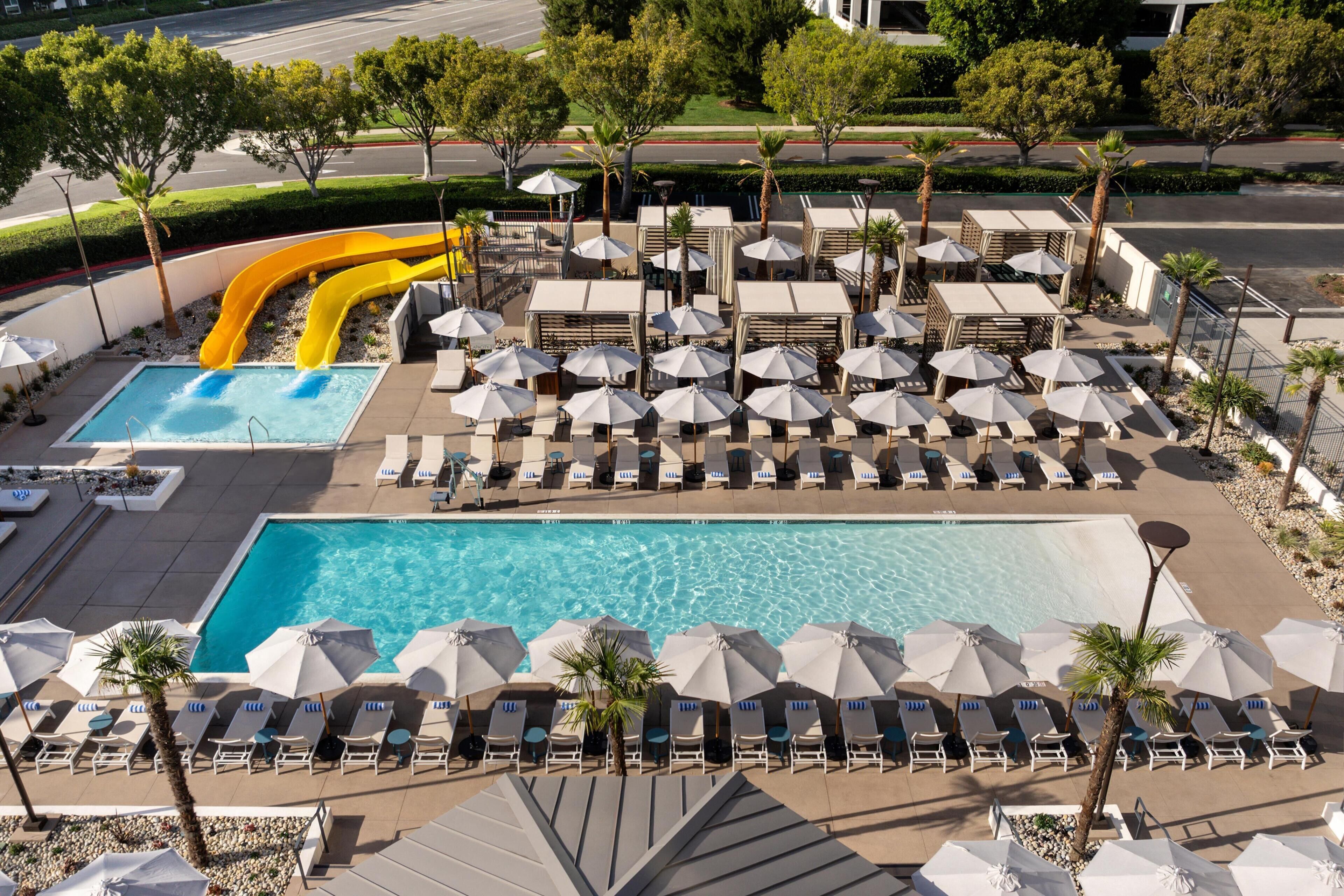Zona de piscina con varias sillas reclinables y sombrillas, una piscina grande y un tobogán de agua.