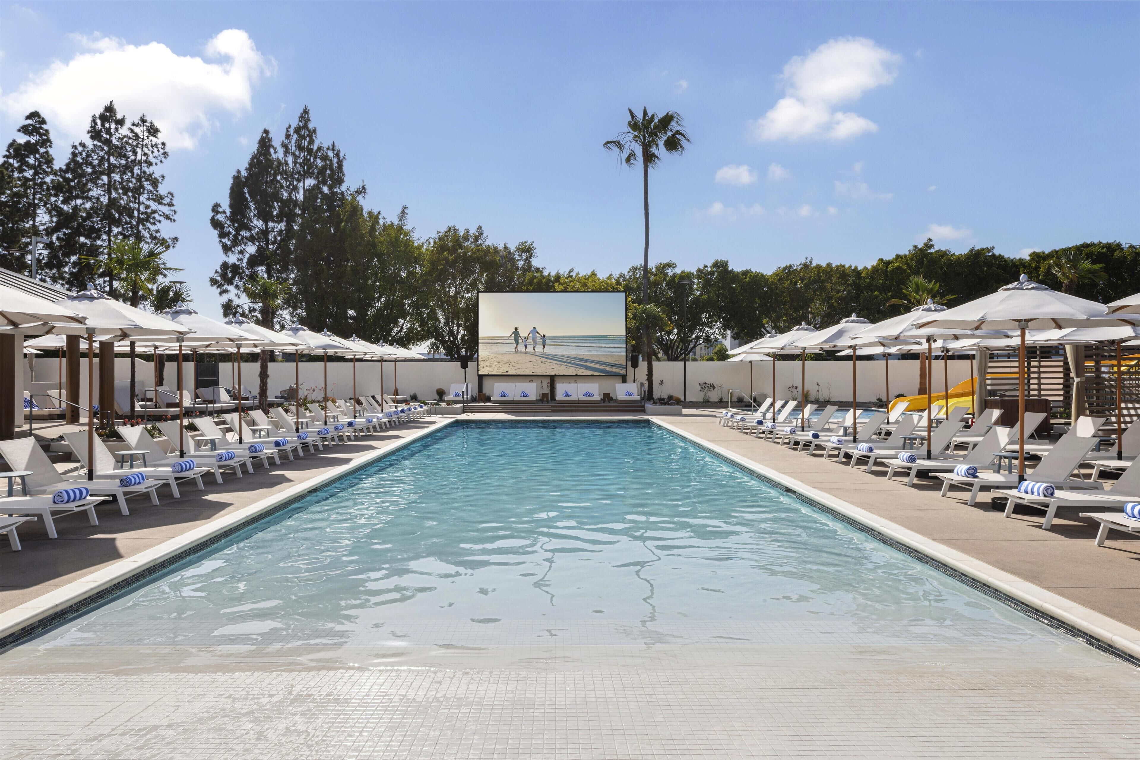 A swimming pool with lounge chairs and umbrellas, surrounded by trees and a clear sky.