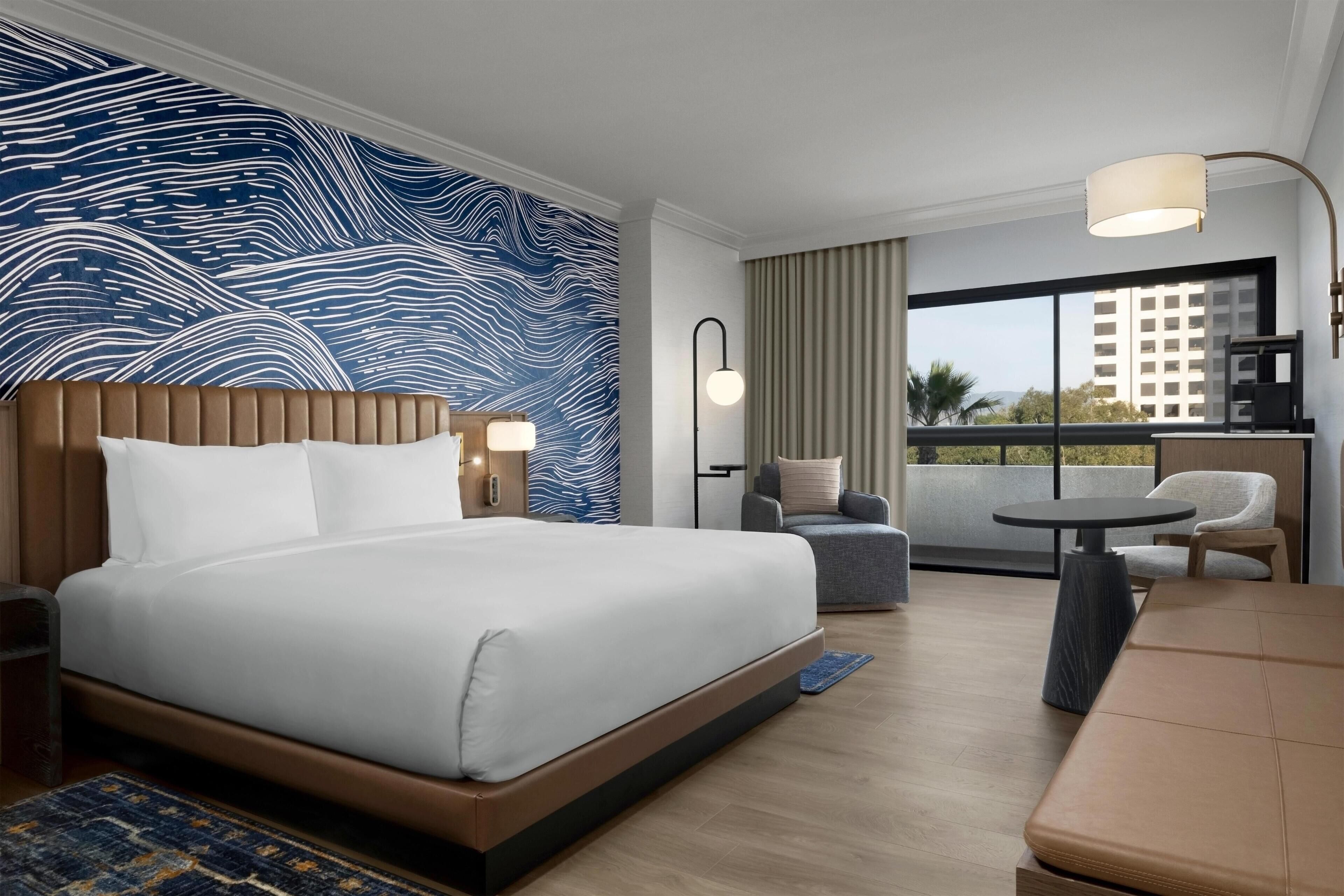 A hotel room with a large bed, a seating area, a balcony with a view, and a modern design.