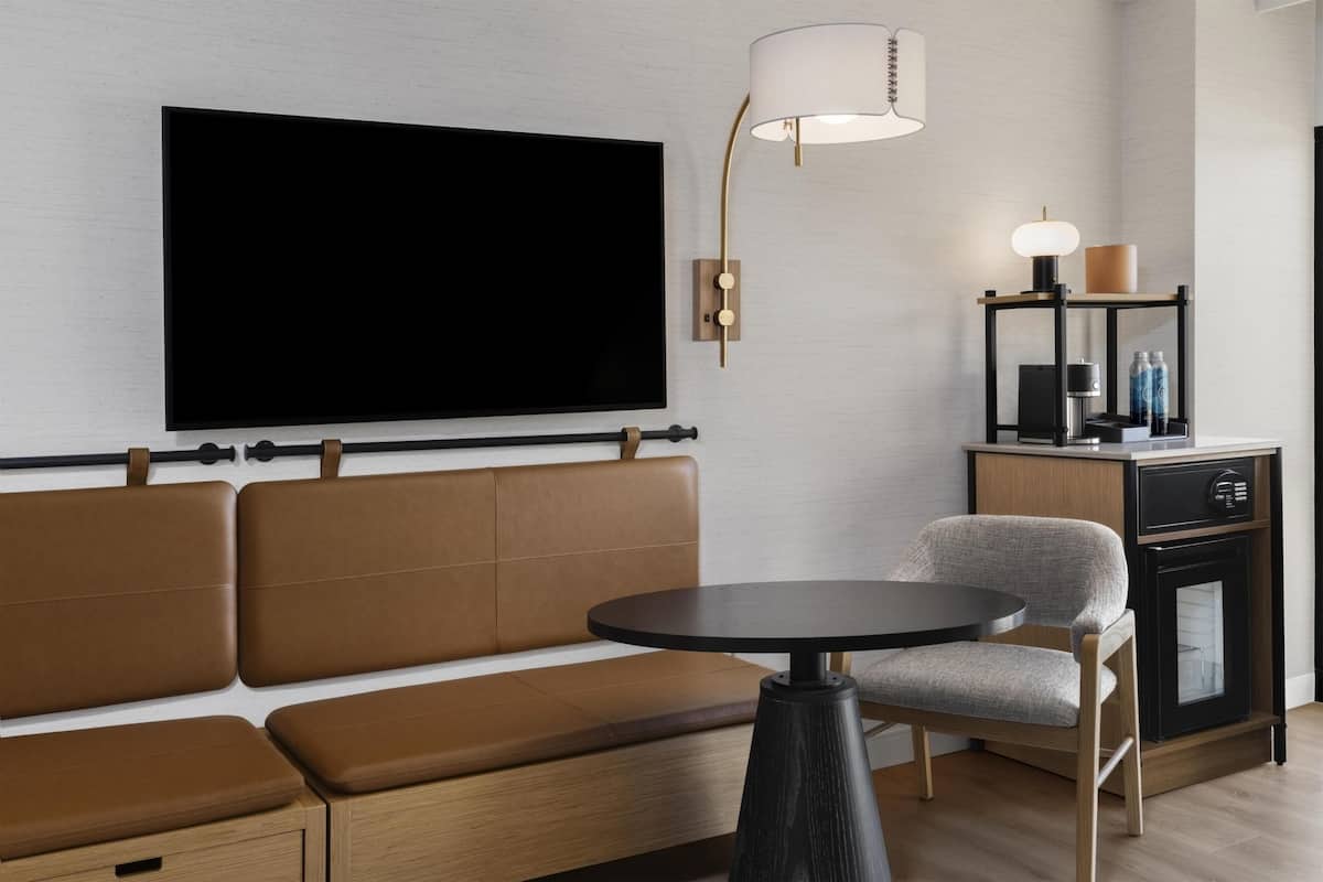 Ein modernes Wohnzimmer mit einem großen Flachbildfernseher, einer Couch, einem runden Tisch, einer Lampe und einer kleinen Vitrine mit Flaschen.