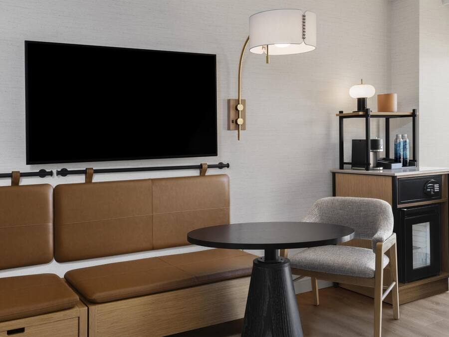 Uma sala de estar moderna com uma televisão de tela plana grande, um sofá, uma mesa redonda, um abajur e um pequeno armário com garrafas.