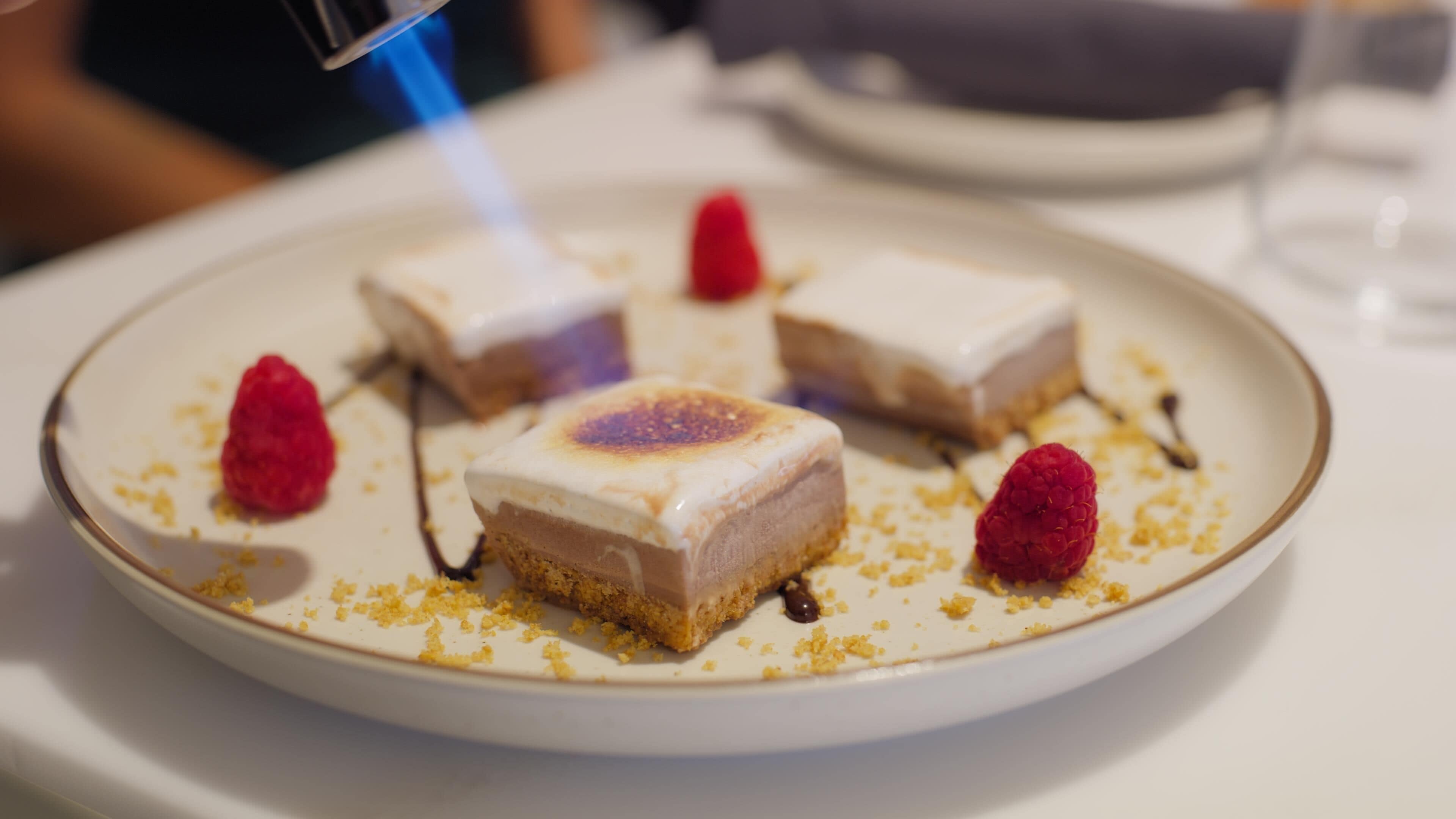 Ein Dessertteller mit einem Dessert, das eine gebrannte Oberfläche hat, umgeben von Himbeeren und Krümeln.