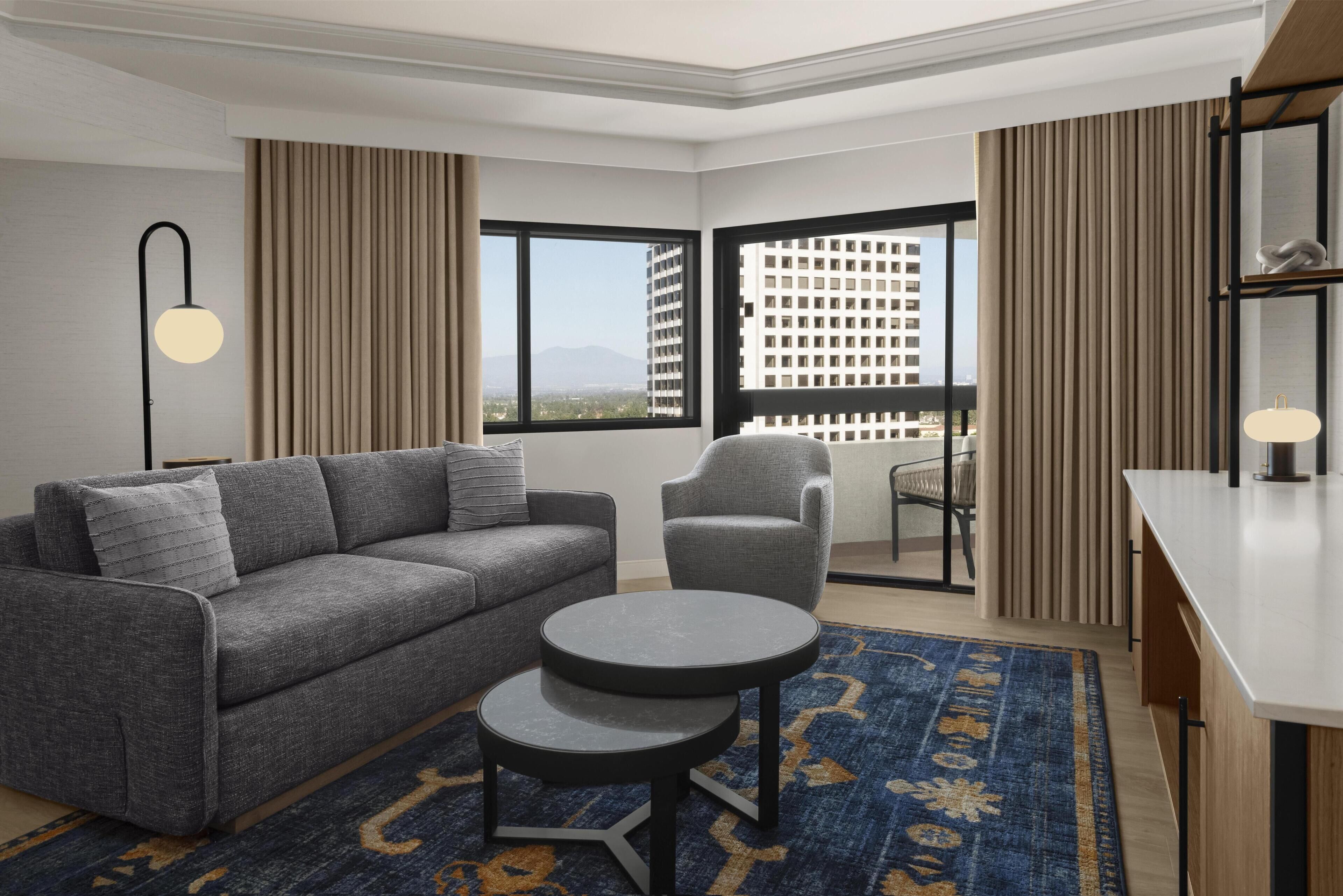 Una habitación de hotel moderna con un sofá gris, una pequeña mesa redonda, una silla, un escritorio y vistas a un paisaje urbano a través de ventanas amplias.