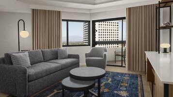 Una habitación de hotel moderna con un sofá gris, una pequeña mesa redonda, una silla, un escritorio y vistas a un paisaje urbano a través de ventanas amplias.