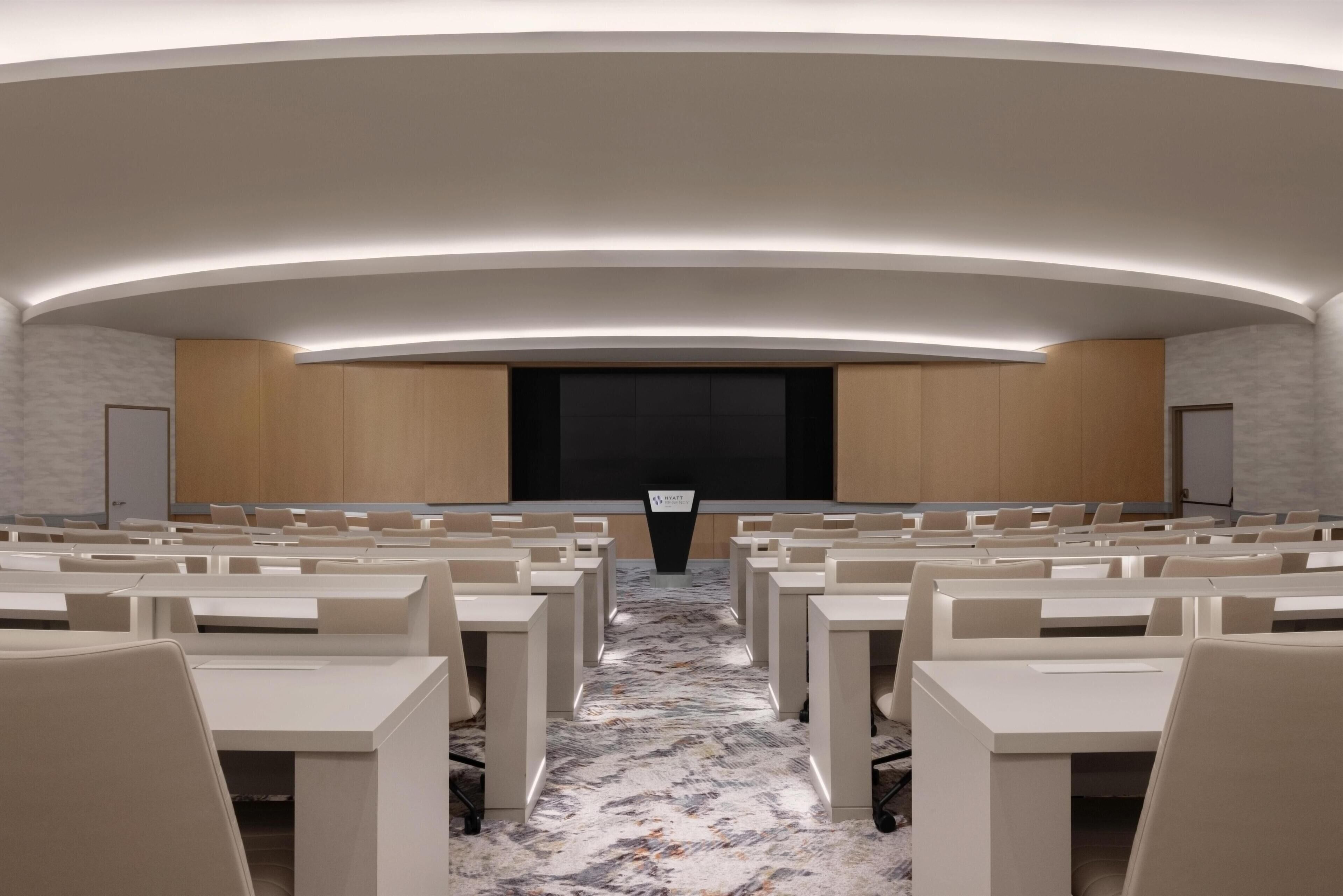 A conference room with rows of tables and chairs, a large screen, and a podium.