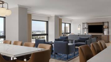 A modern hotel room with a dining area, living room, large windows, and a flat-screen TV.