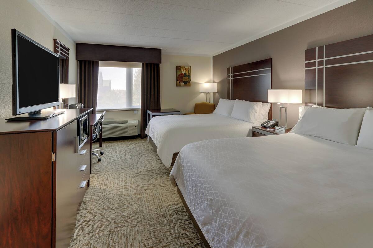 A hotel room with two beds, a desk with a flat-screen TV, a desk lamp, a chair, and a window with curtains.