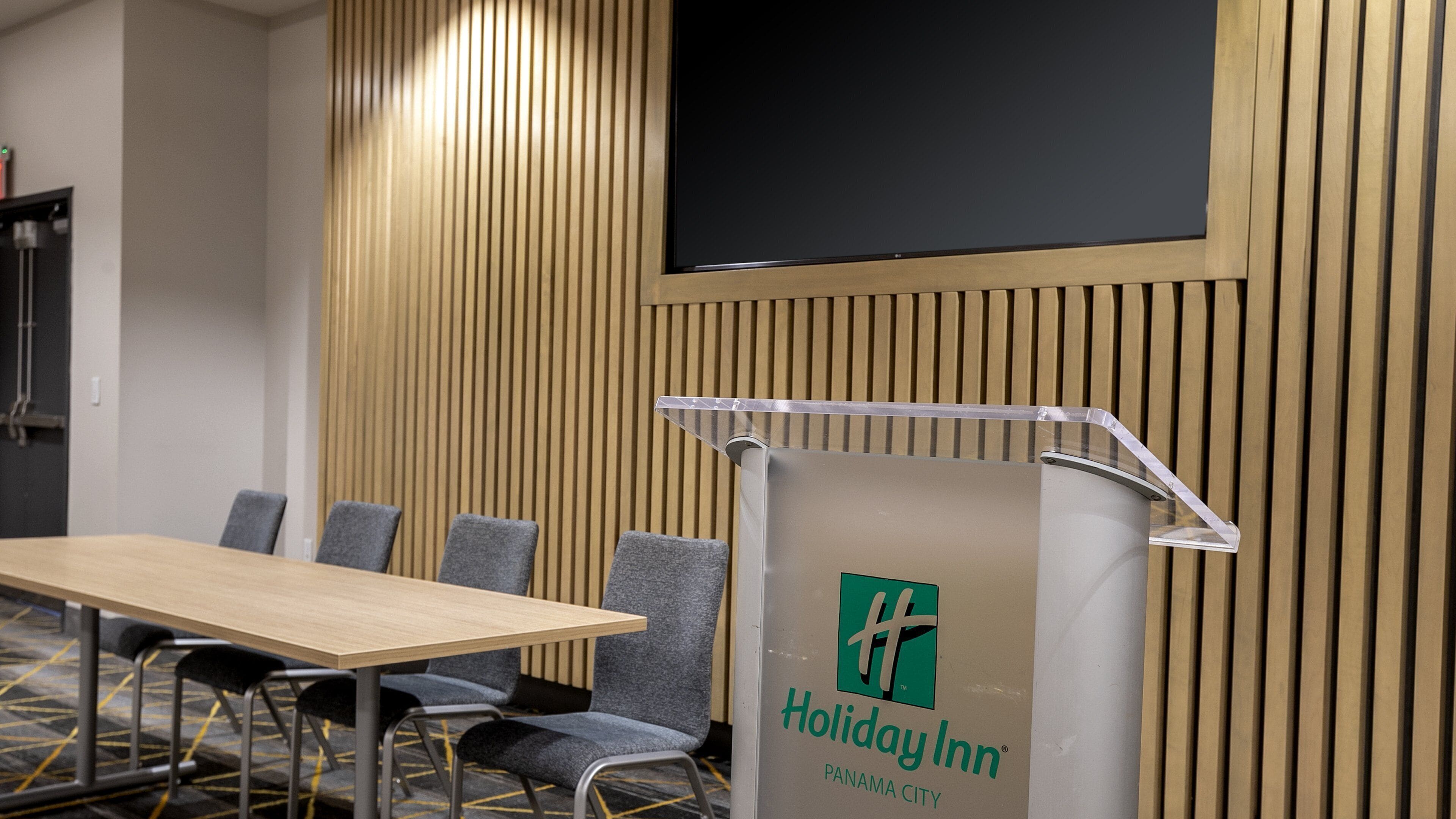 Un salón de conferencias con pared de paneles de madera, un atril con el logotipo de Holiday Inn y una pantalla negra en la parte superior.