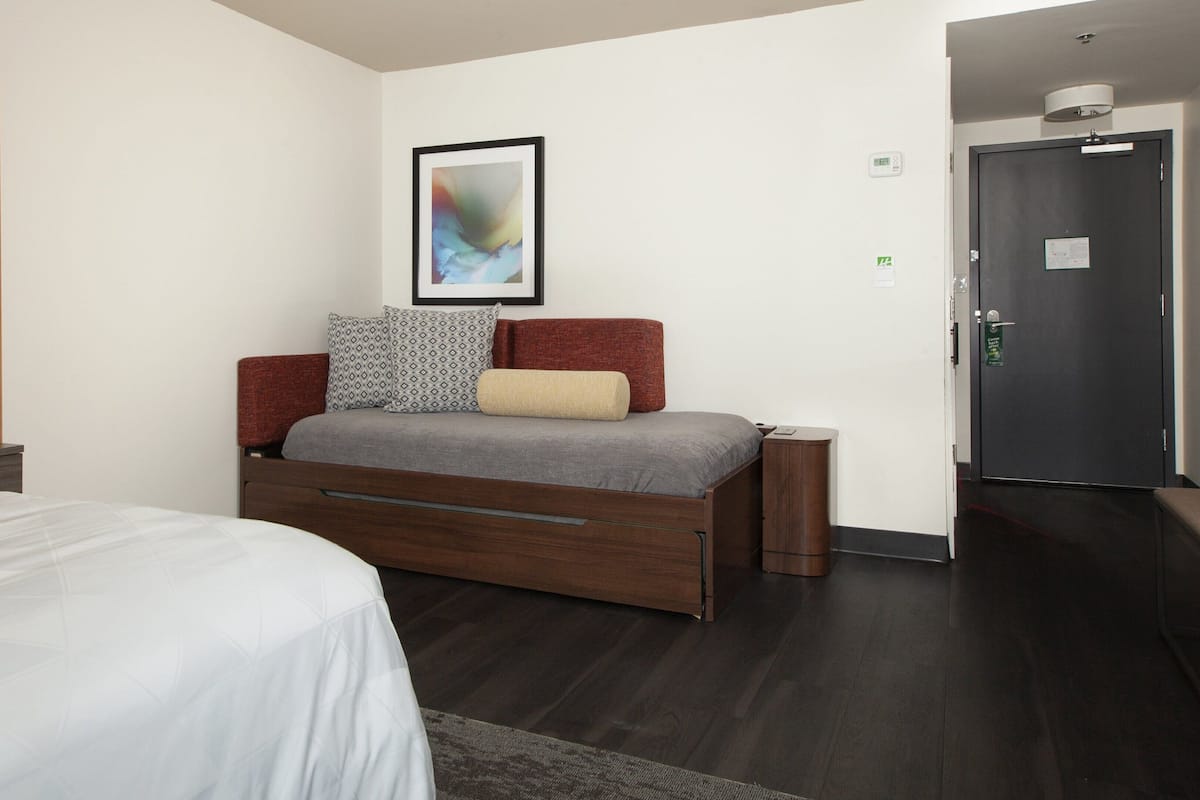 Una habitación de hotel moderna con una cama, un escritorio, una silla, un cuadro abstracto enmarcado, una puerta y un termostato.