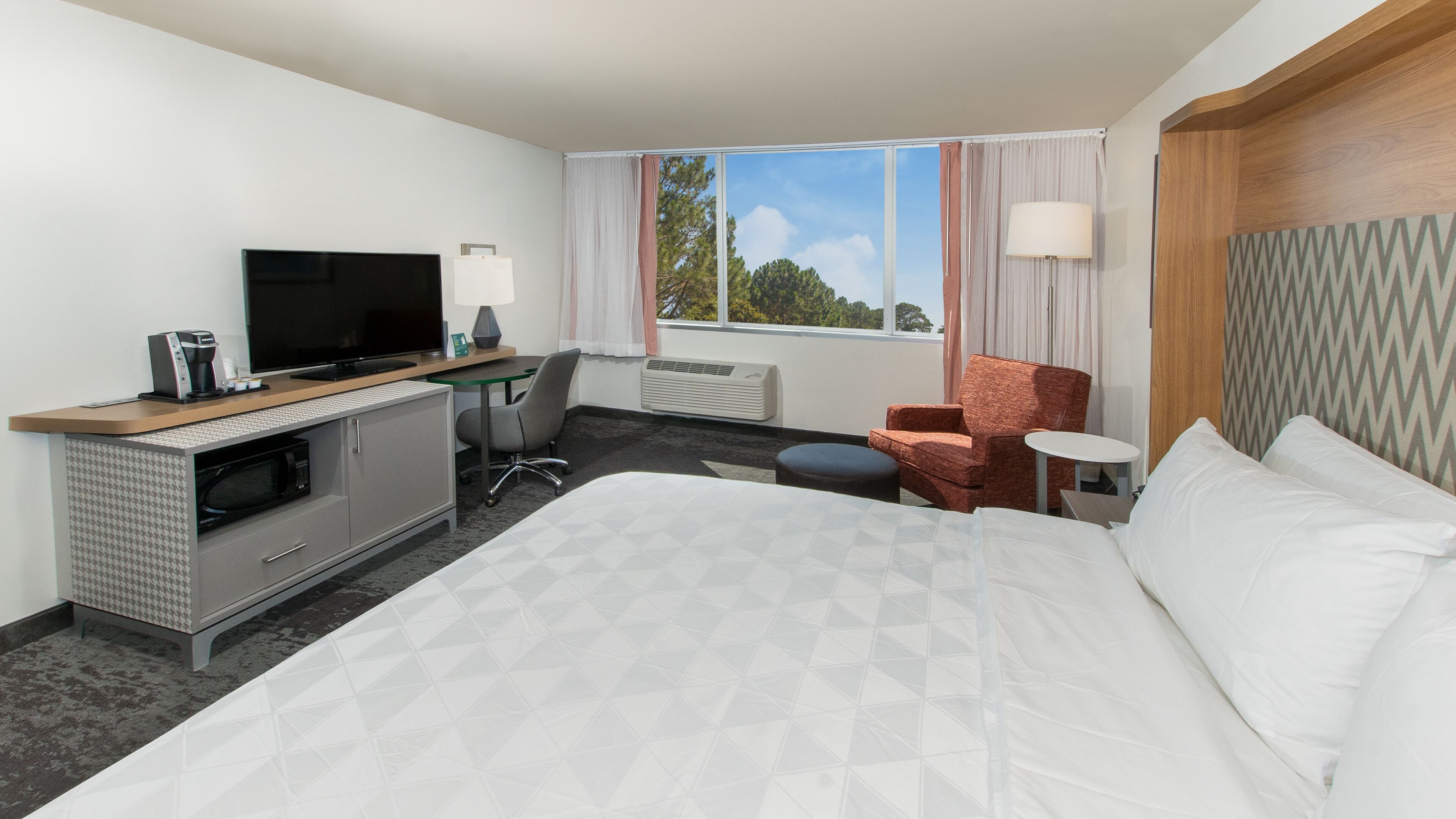 Une chambre d’hôtel comprenant un lit, un bureau avec une télévision, une chaise, une petite table et une fenêtre avec des rideaux.