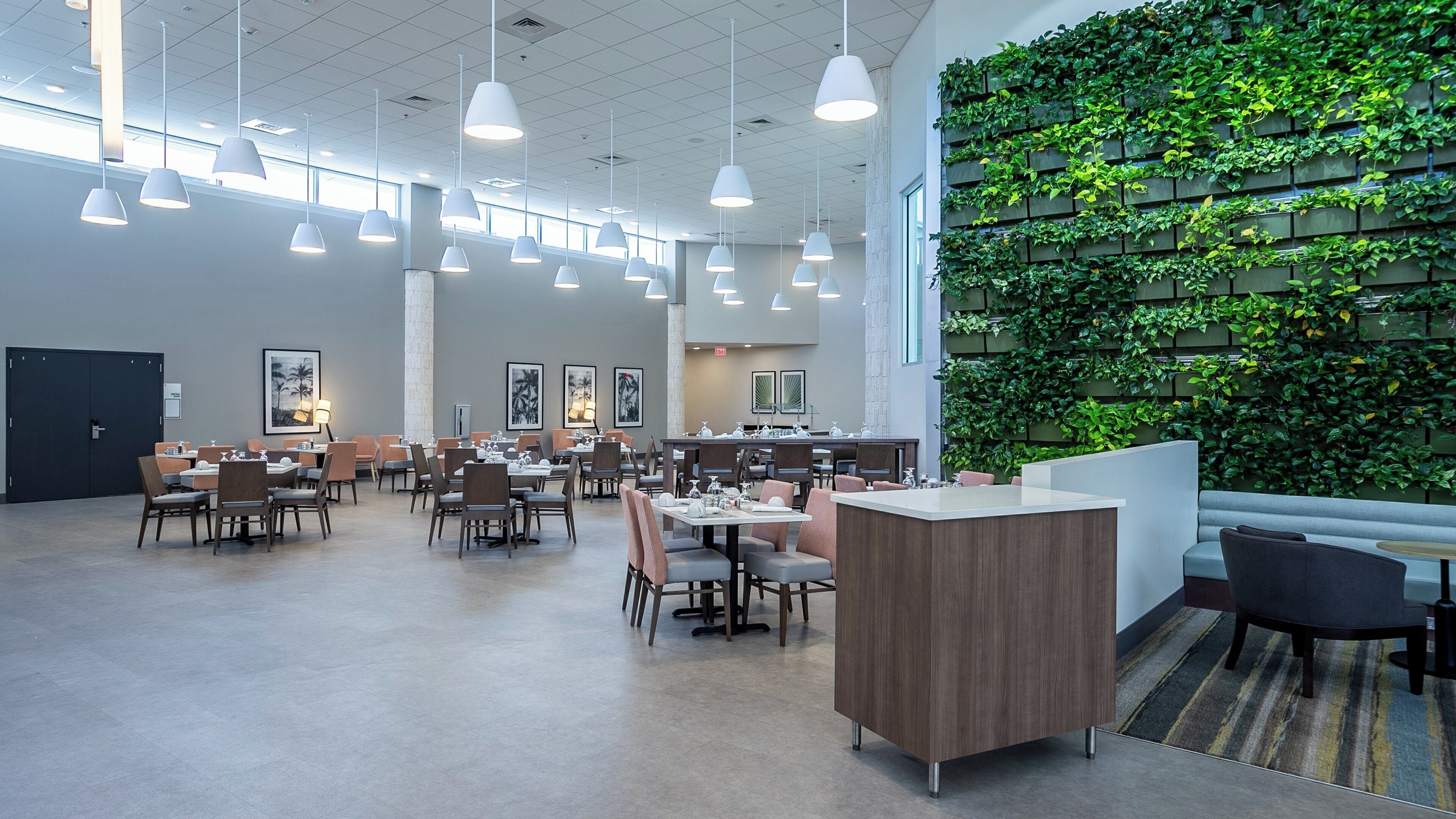 A spacious dining area with multiple tables and chairs, pendant lighting, and a green vertical garden wall.