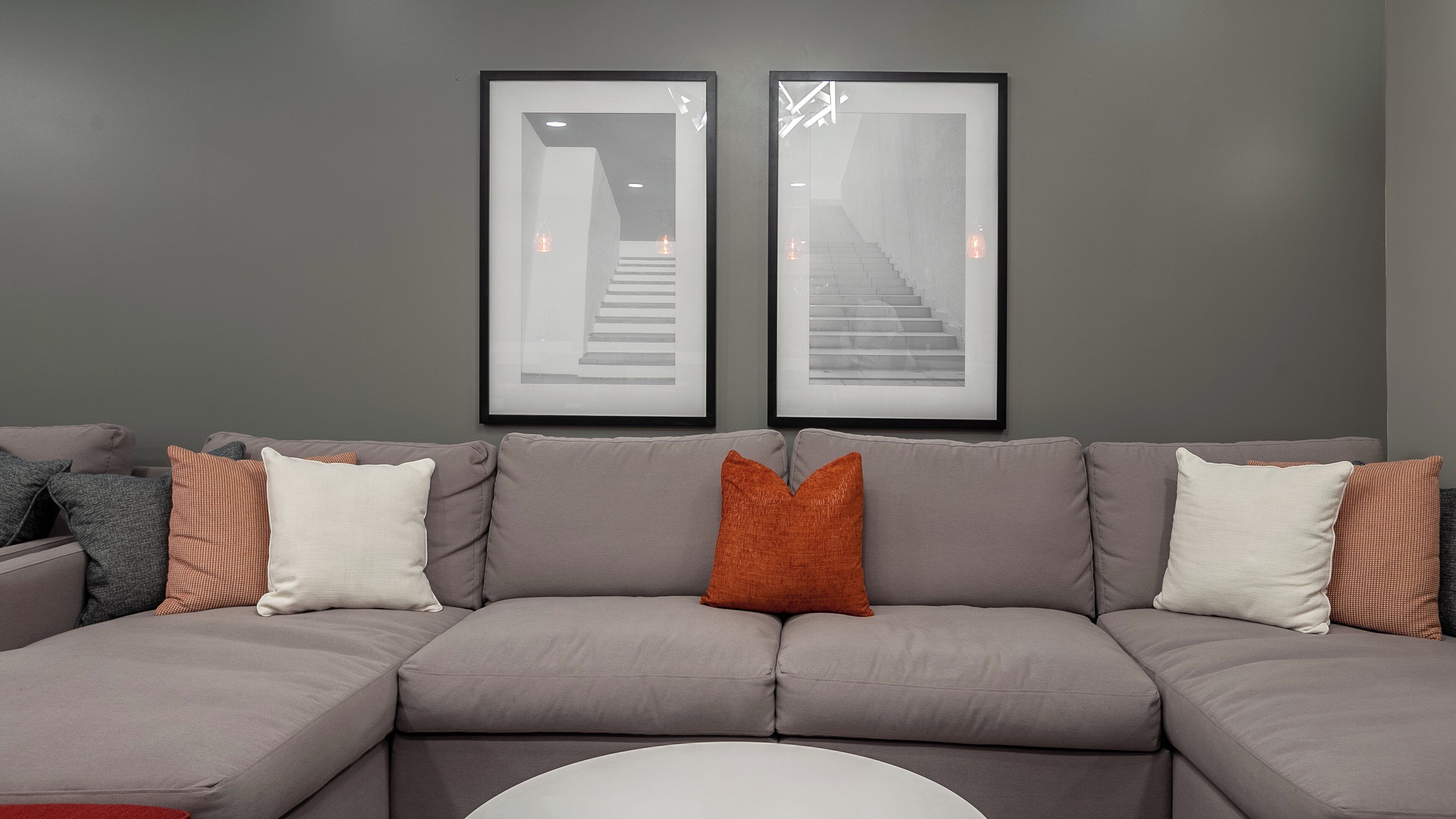 Ein graues Ecksofa mit vielen Kissen, ein weißer Couchtisch und zwei gerahmte Bilder von Treppen an der Wand.
