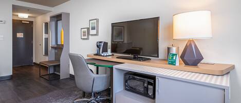 A hotel room with a desk, chair, computer, lamp, and microwave.
