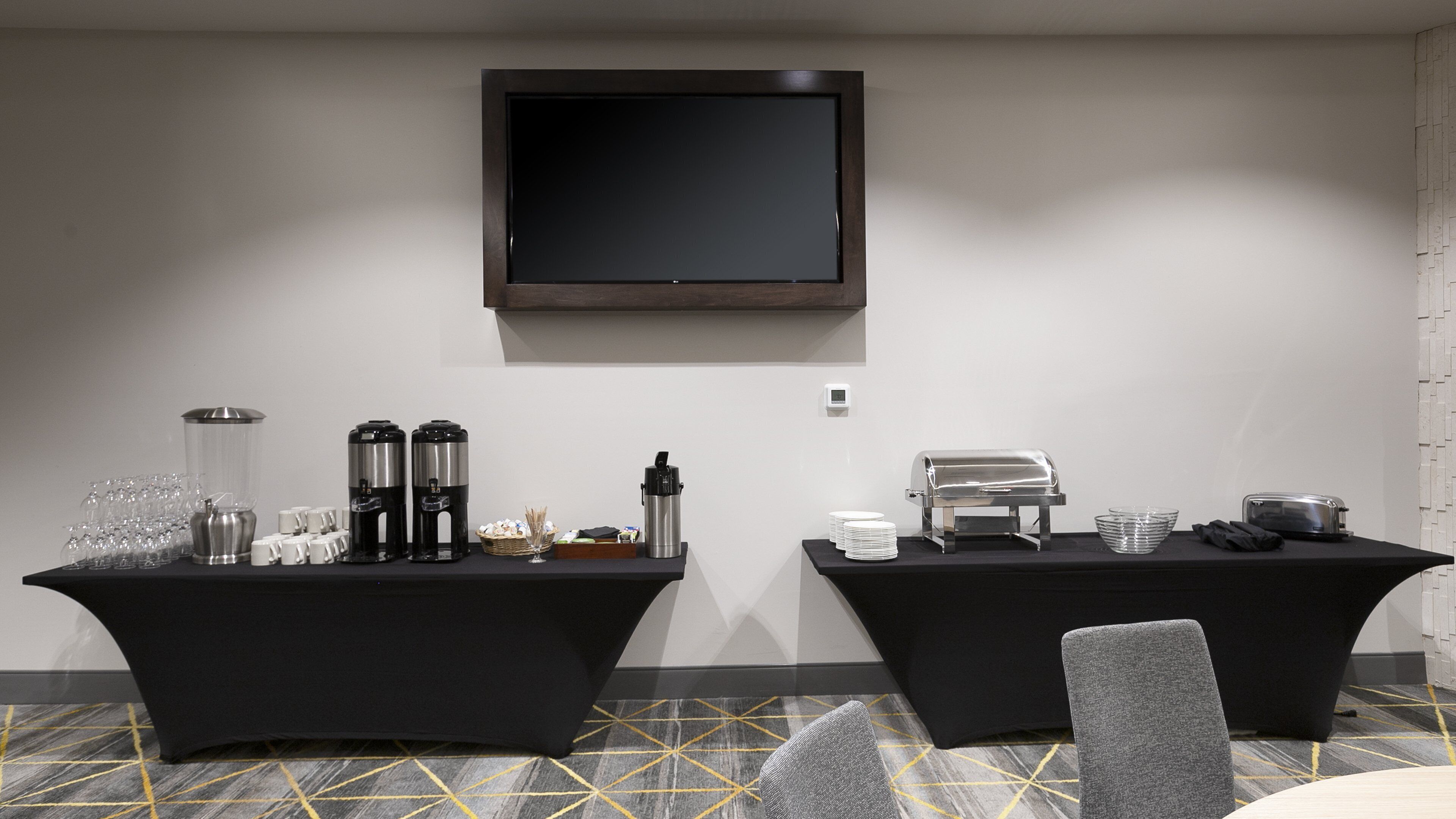 Un salón de conferencias con dos mesas, cada una con cafeteras, tazas y un tostador. Un televisor de pantalla plana está montado en la pared sobre las mesas.