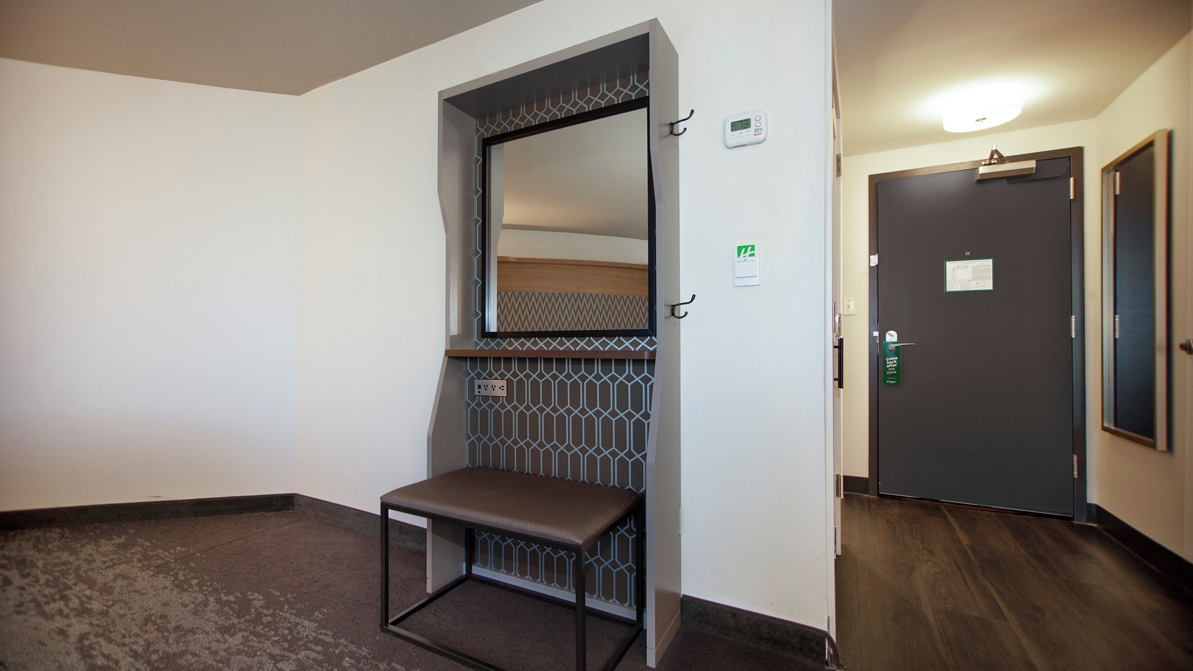 Een hotelkamer met een bankje, een deur met slot, een wandthermostaat en een lamp.