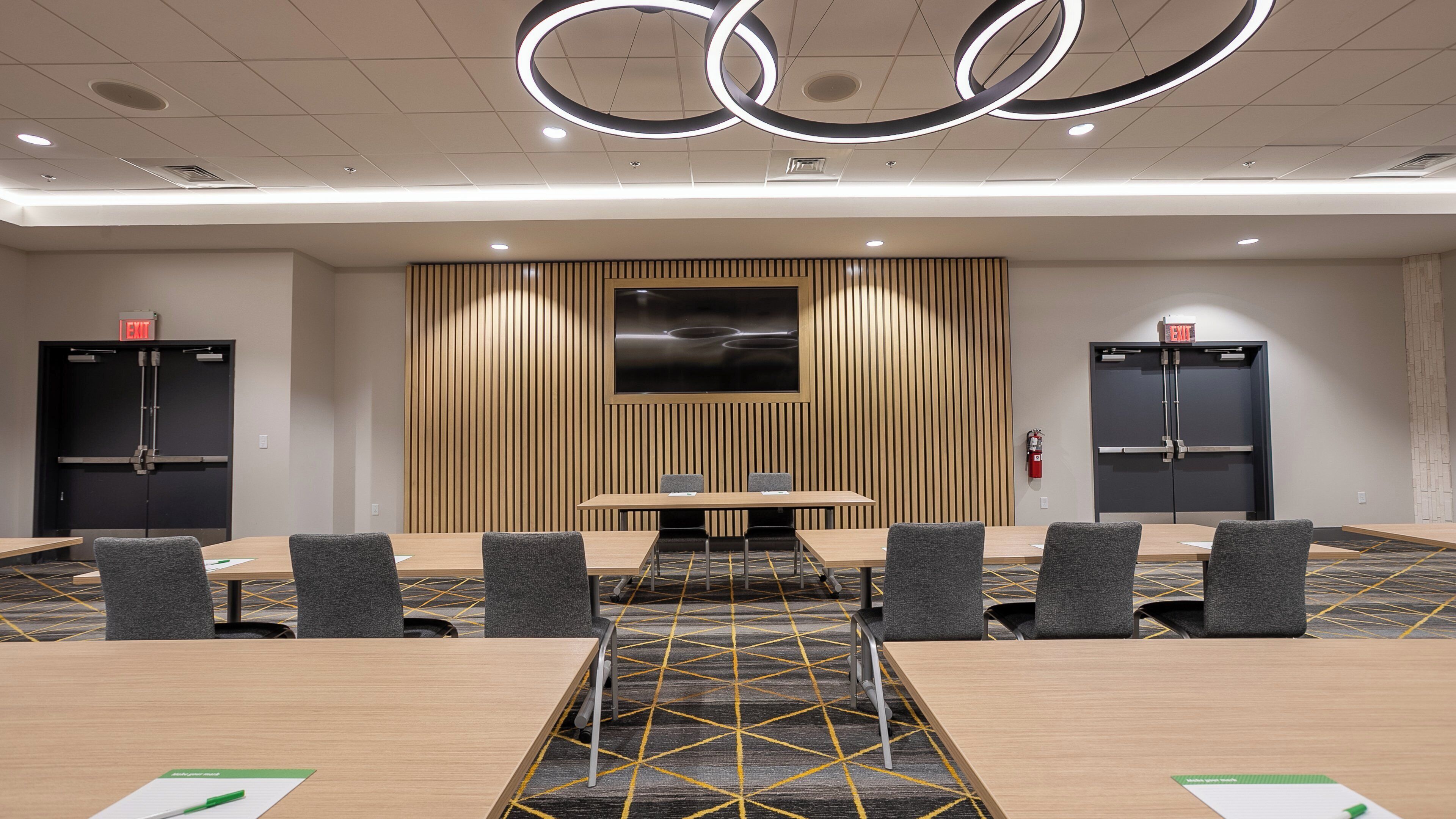 A conference room with a long table, six chairs, a large screen, and two black cabinets with red exit signs.