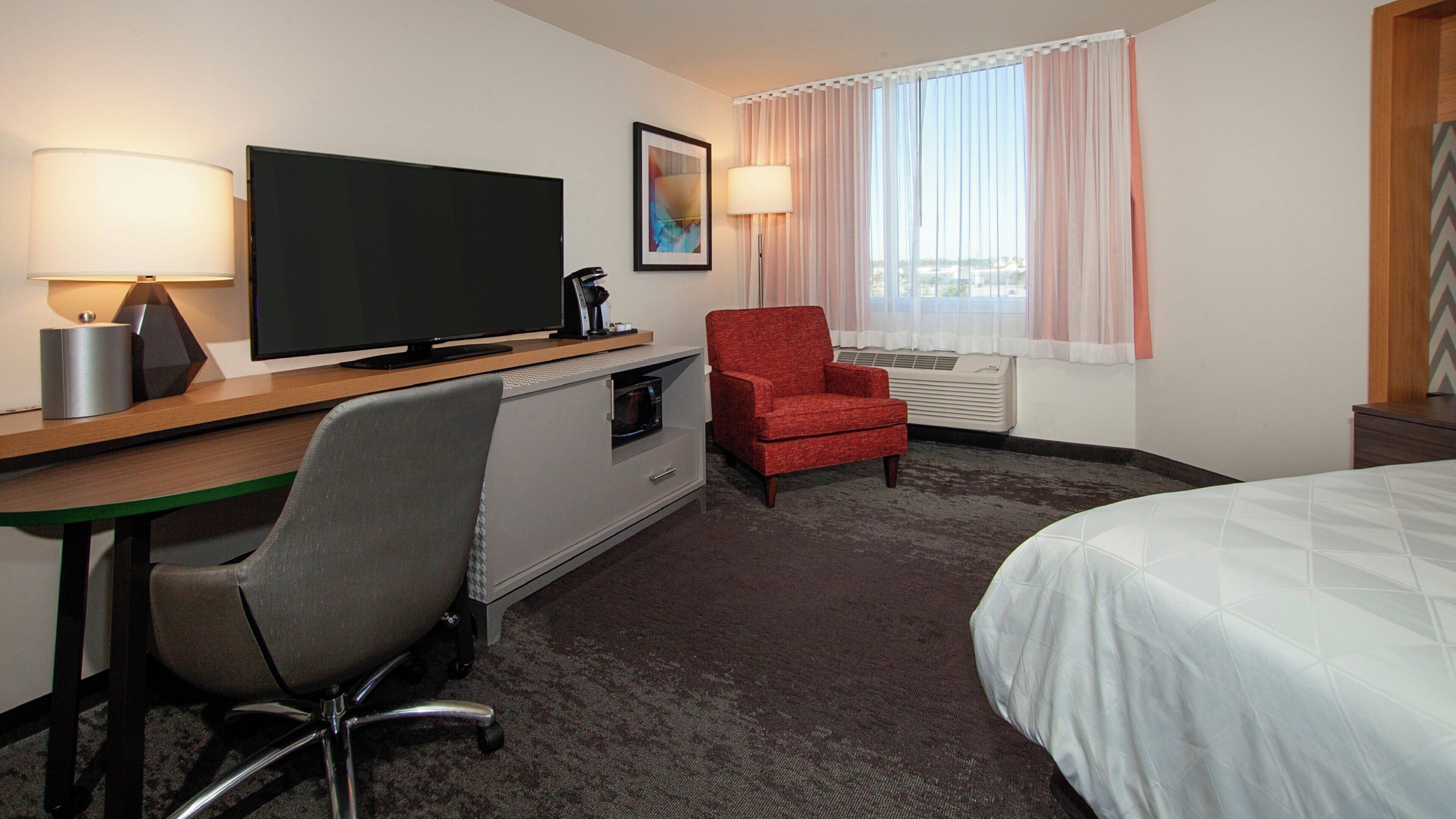 A hotel room with a bed, a desk with a television, a red armchair, a microwave, and a window with curtains.