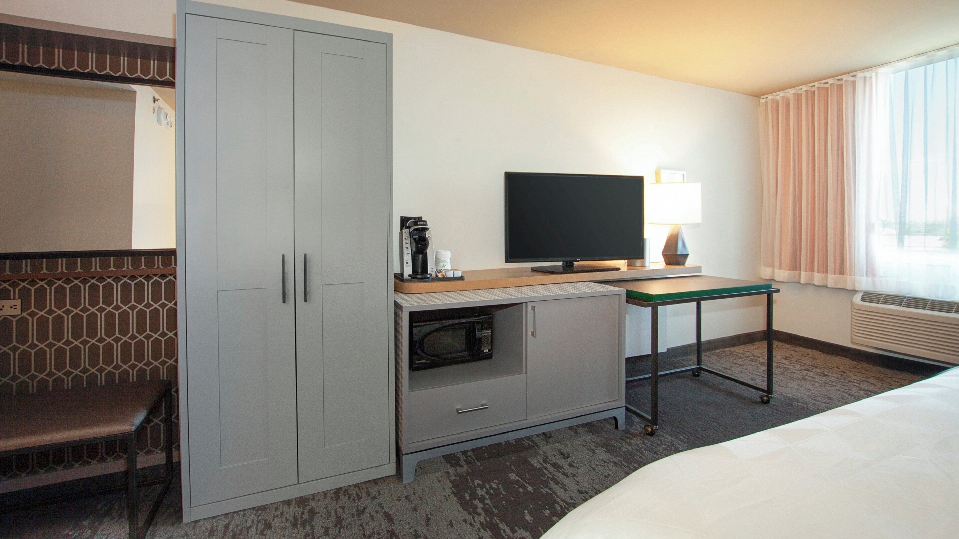 A hotel room with a bed, a desk with a flat-screen TV, a microwave, and a seating area with a patterned wall.