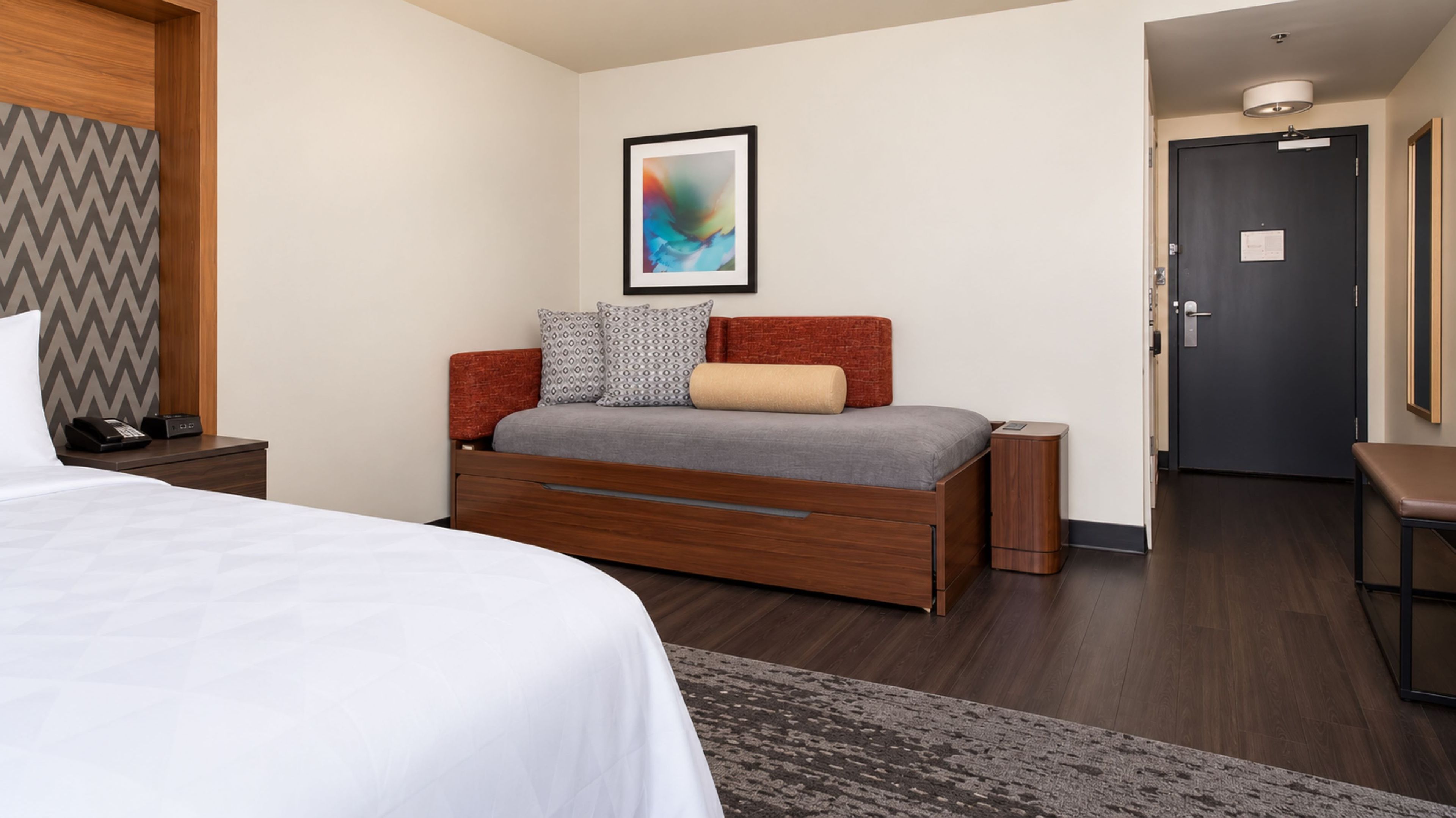 Quarto de hotel com cama, mesa de cabeceira, pequena mesa de cabeceira com abajur, obra de arte pendurada na parede e uma porta.
