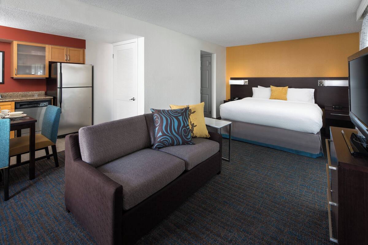 A hotel room with a sofa, a bed, a TV, a desk with a chair, and a kitchen area with a refrigerator.