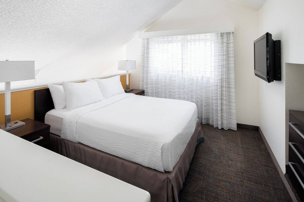 A hotel room with a neatly made bed, a flat-screen TV mounted on the wall, and a window with sheer curtains.