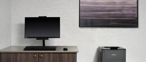 A modern office setup with a desktop computer, keyboard, and mouse on a wooden desk. A Brother printer is on the floor beside the desk. A framed abstract painting is on the wall.