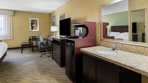 Quarto de hotel com cama, escrivaninha, televisão, micro-ondas, pia com espelho e uma cadeira.
