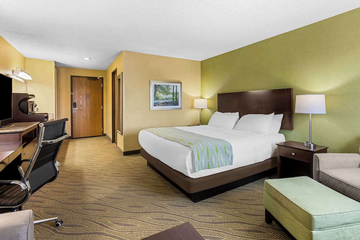 A hotel room with a large bed, a desk with a chair, a small table, and a lamp.