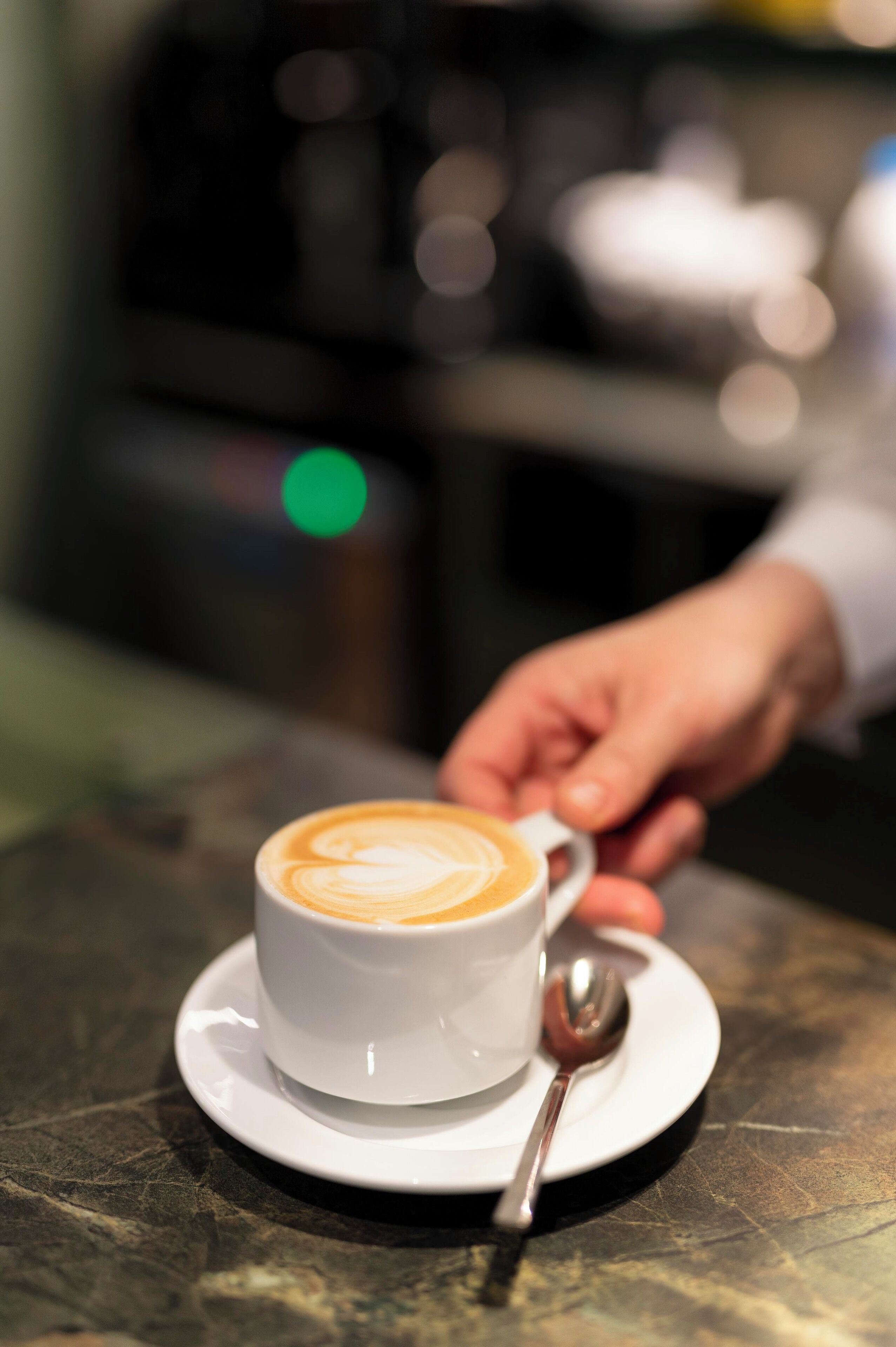 Una taza de café con arte de latte sobre un plato, una cuchara y una mano alcanzando la taza.