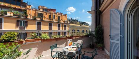A terrace with a table and chairs, surrounded by potted plants and overlooking a street with buildings.
