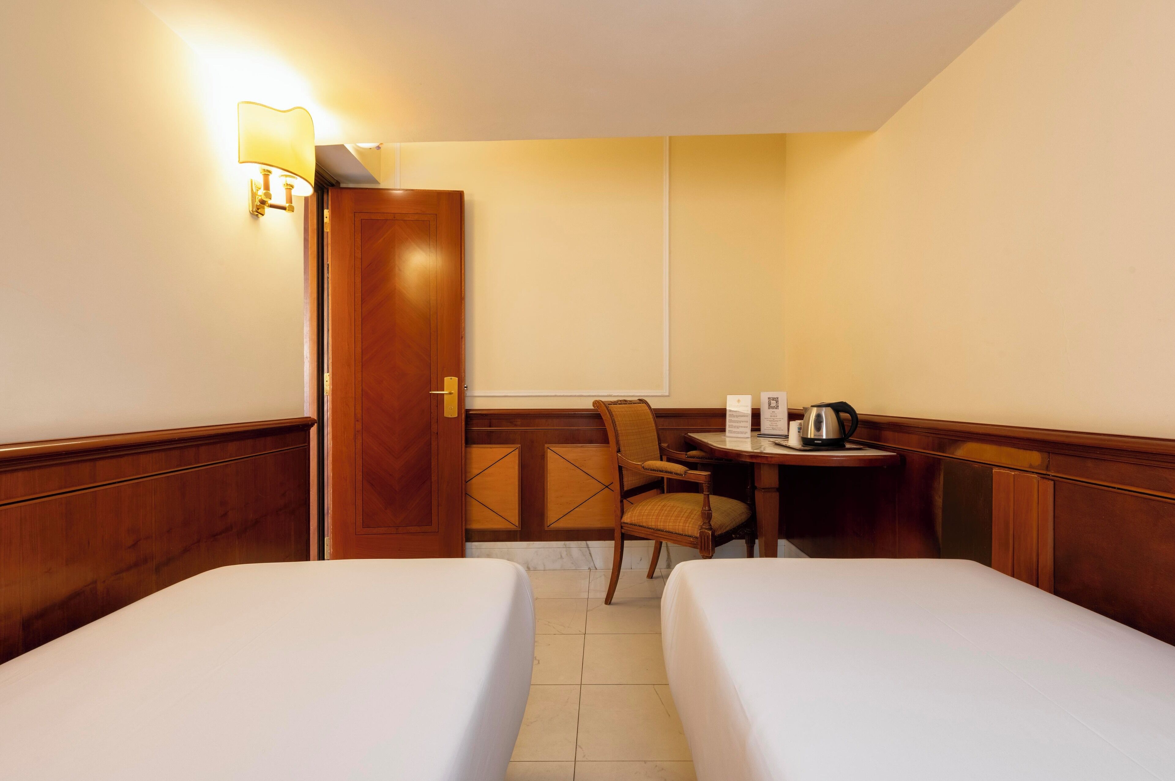 A hotel room with two beds, a wooden door, a small table with a kettle, and a chair.
