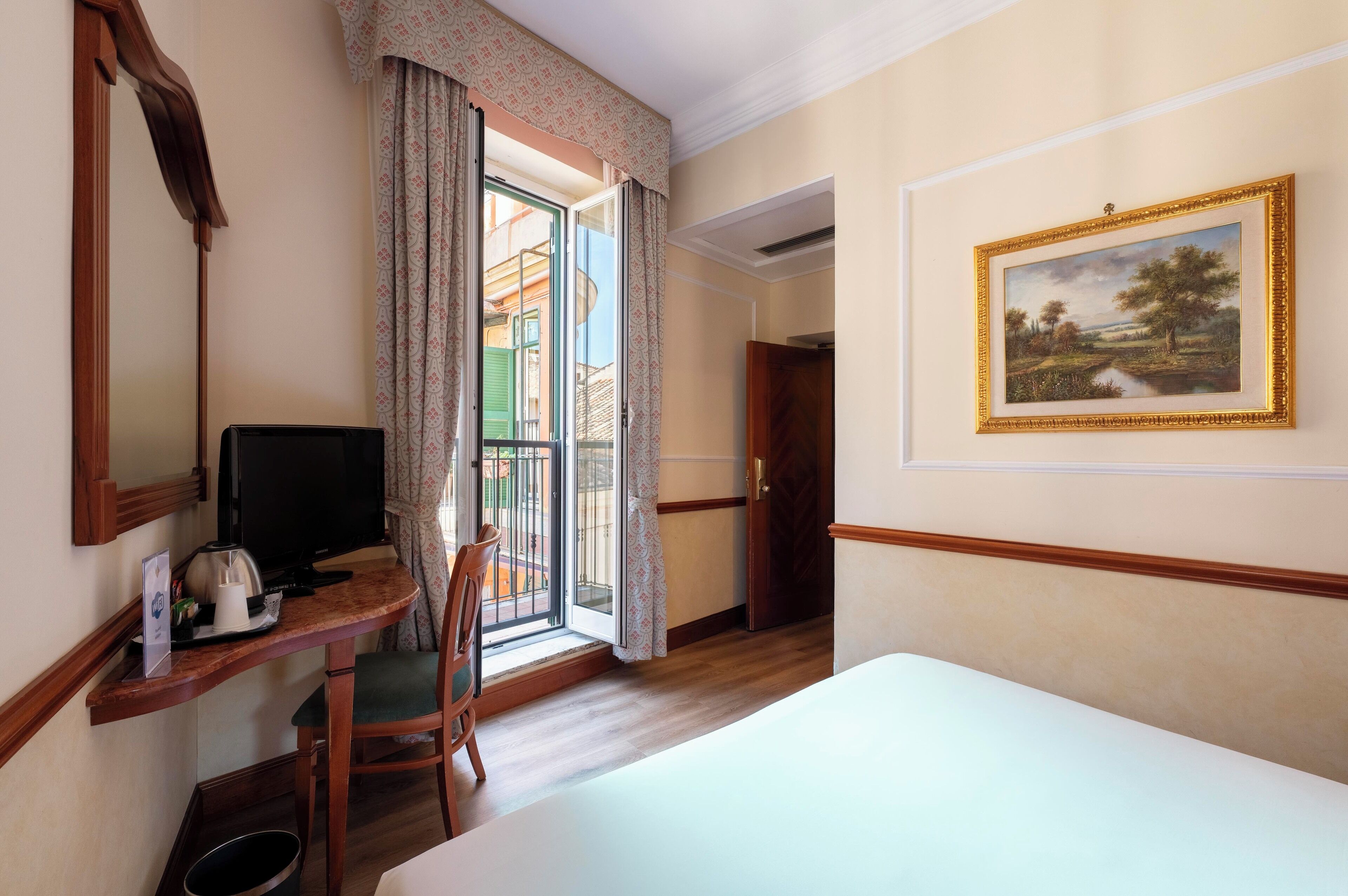 Habitació d'hotel amb un llit, una taula de treball amb un ordinador, una cadira, una finestra amb cortines i un quadre al mur.