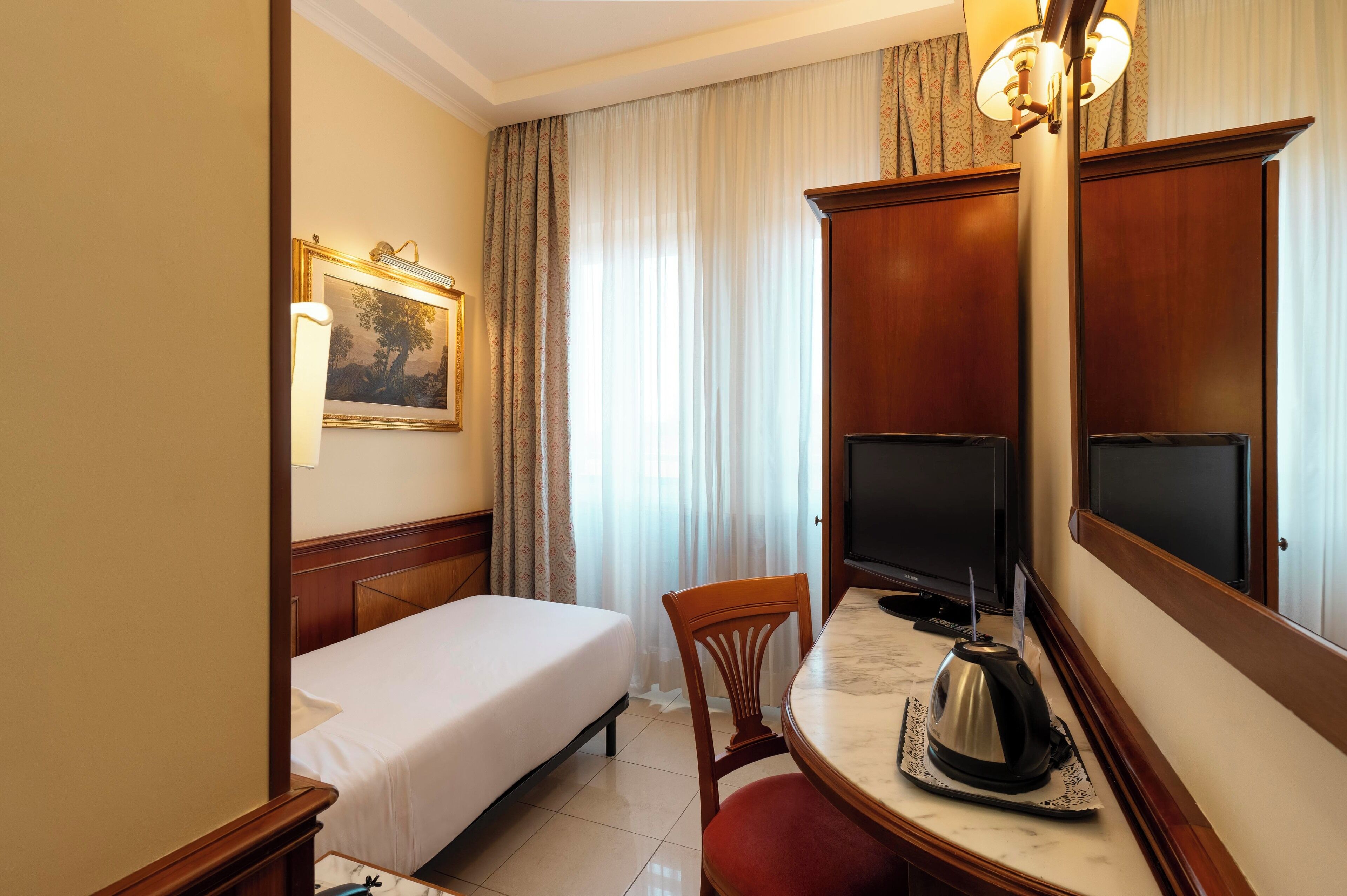 Une chambre d’hôtel avec un lit simple, un bureau avec une bouilloire, une télévision et un grand miroir.