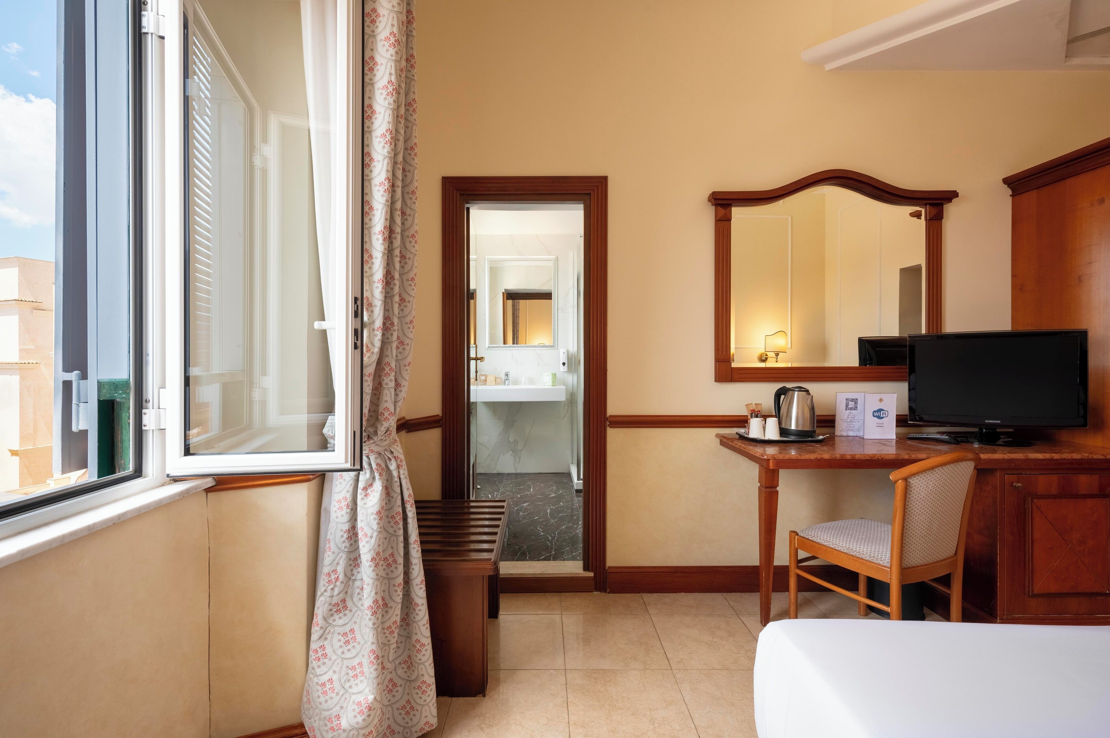 A hotel room with a bed, a desk with a chair, a television, a mirror, and a view of the bathroom through an open door.