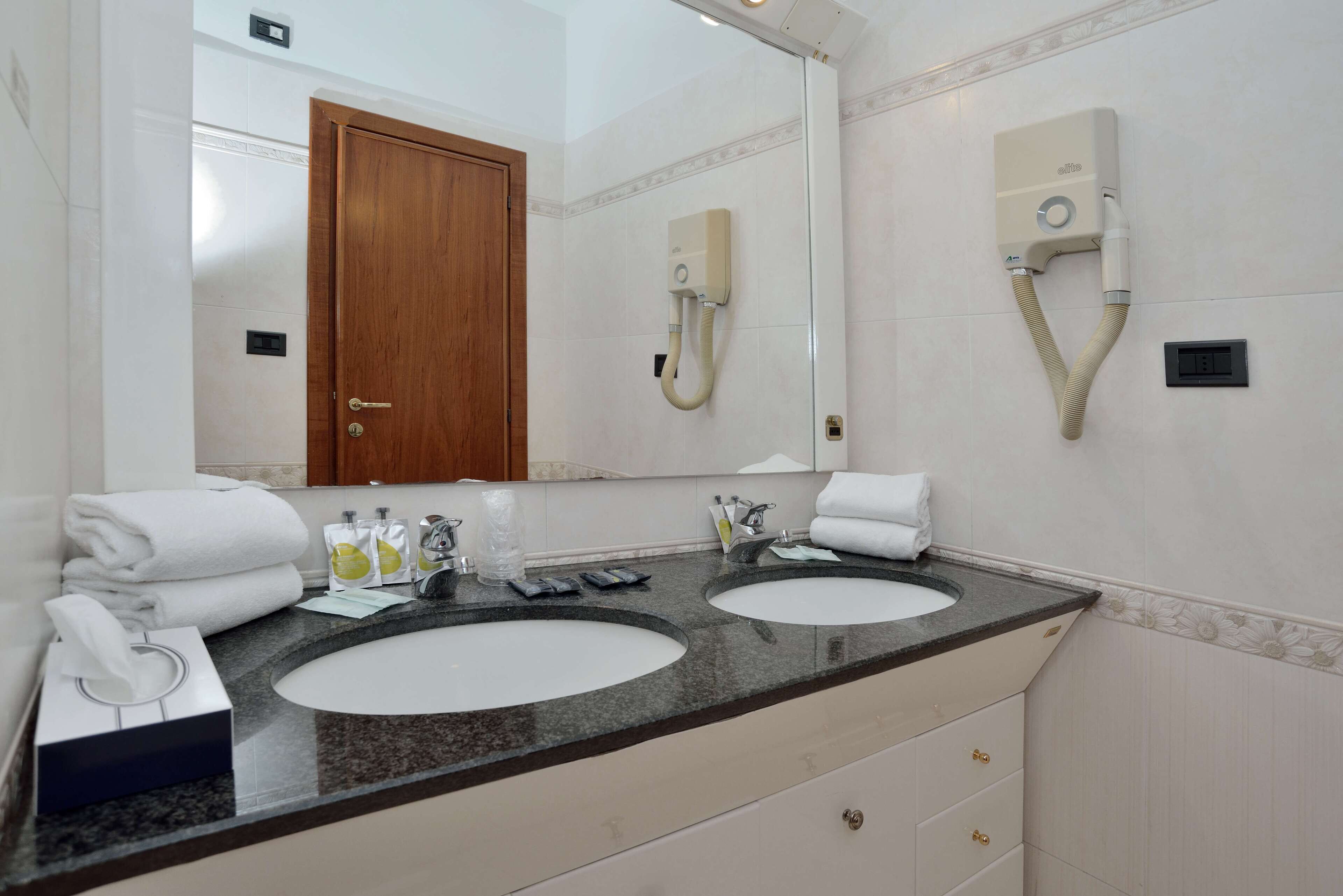 A bathroom with two sinks, a large mirror, a hairdryer mounted on the wall, and neatly folded towels on the counter.