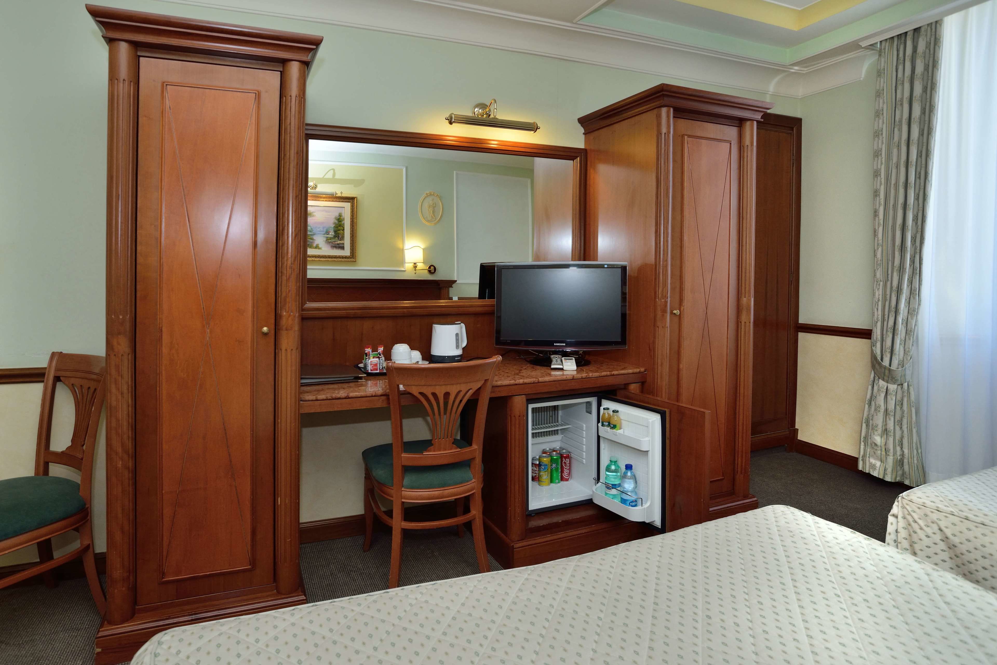 A hotel room with a wooden desk, a television, a small refrigerator, a chair, and a bed.