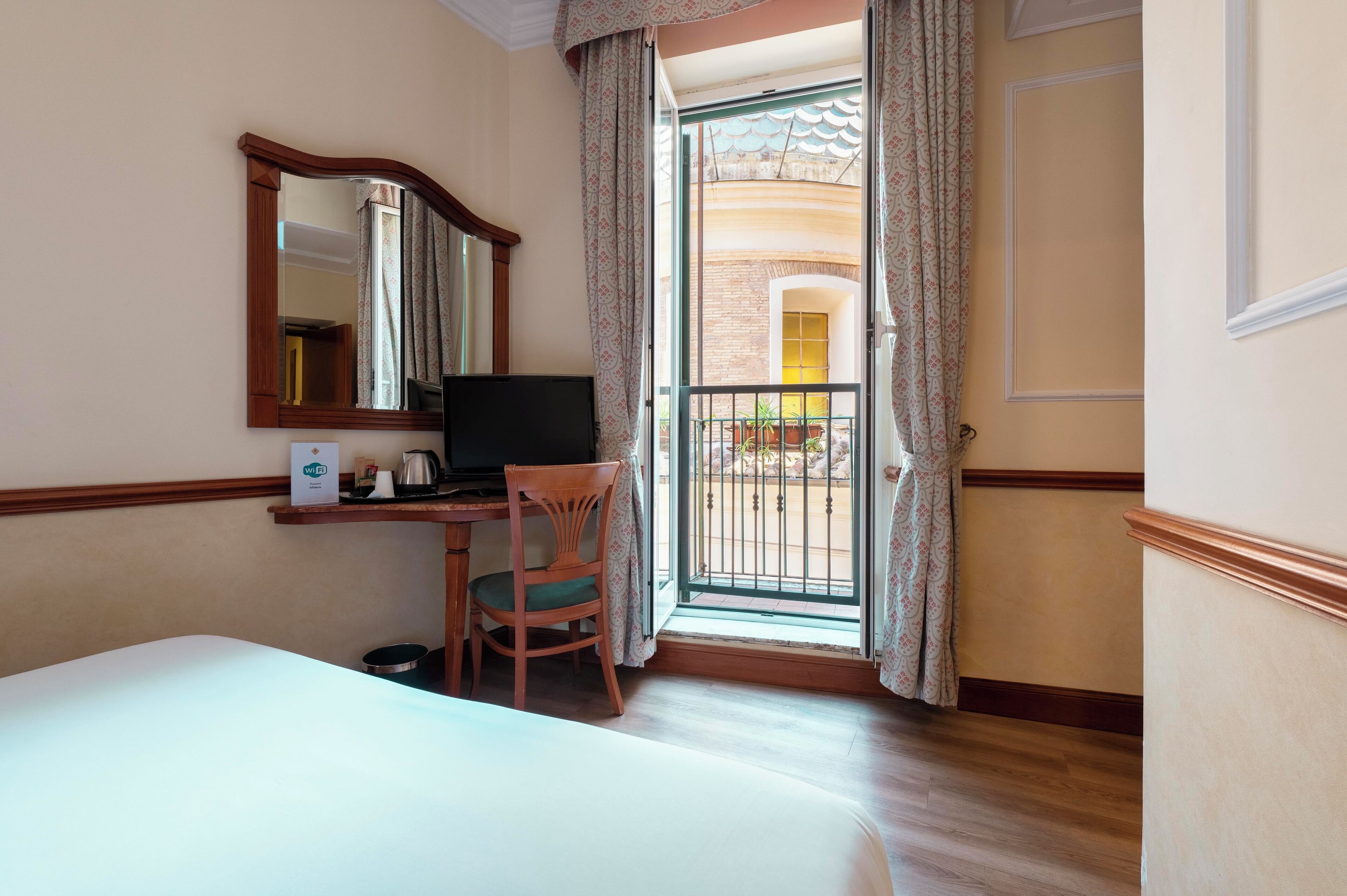 A hotel room with a bed, a desk with a computer, a chair, a TV, and a balcony with a view of the outside.