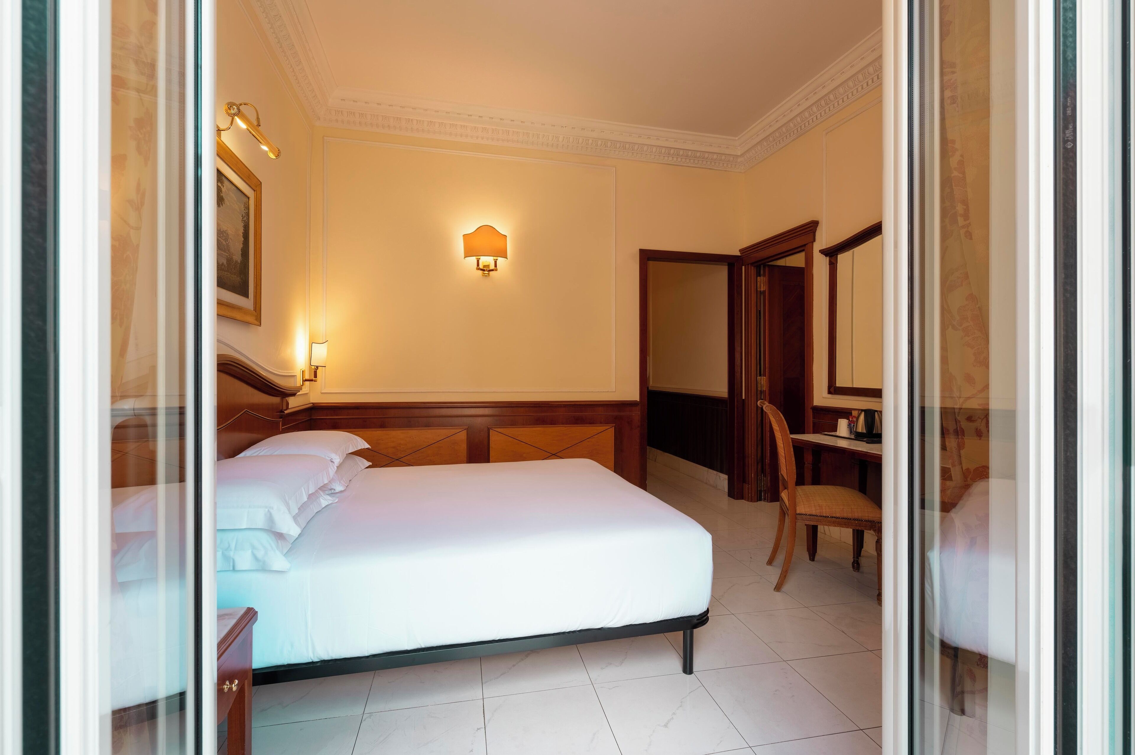 A hotel room with a large bed, a wooden headboard, a desk with a chair, a wall-mounted lamp, and a mirror.