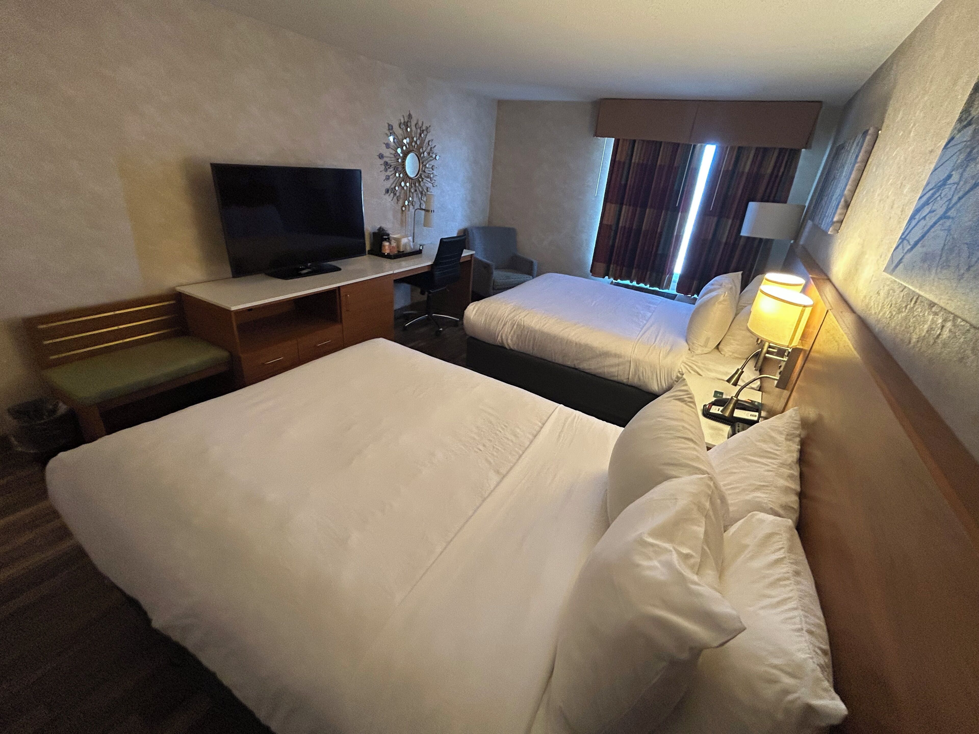 A hotel room with two beds, a desk with a TV, a lamp, and a decorative mirror.