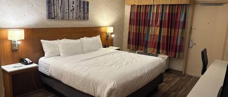 Habitación de hotel con una cama grande, cabecera de madera, mesita de noche con teléfono y lámpara, un cuadro en la pared y una ventana con cortinas.