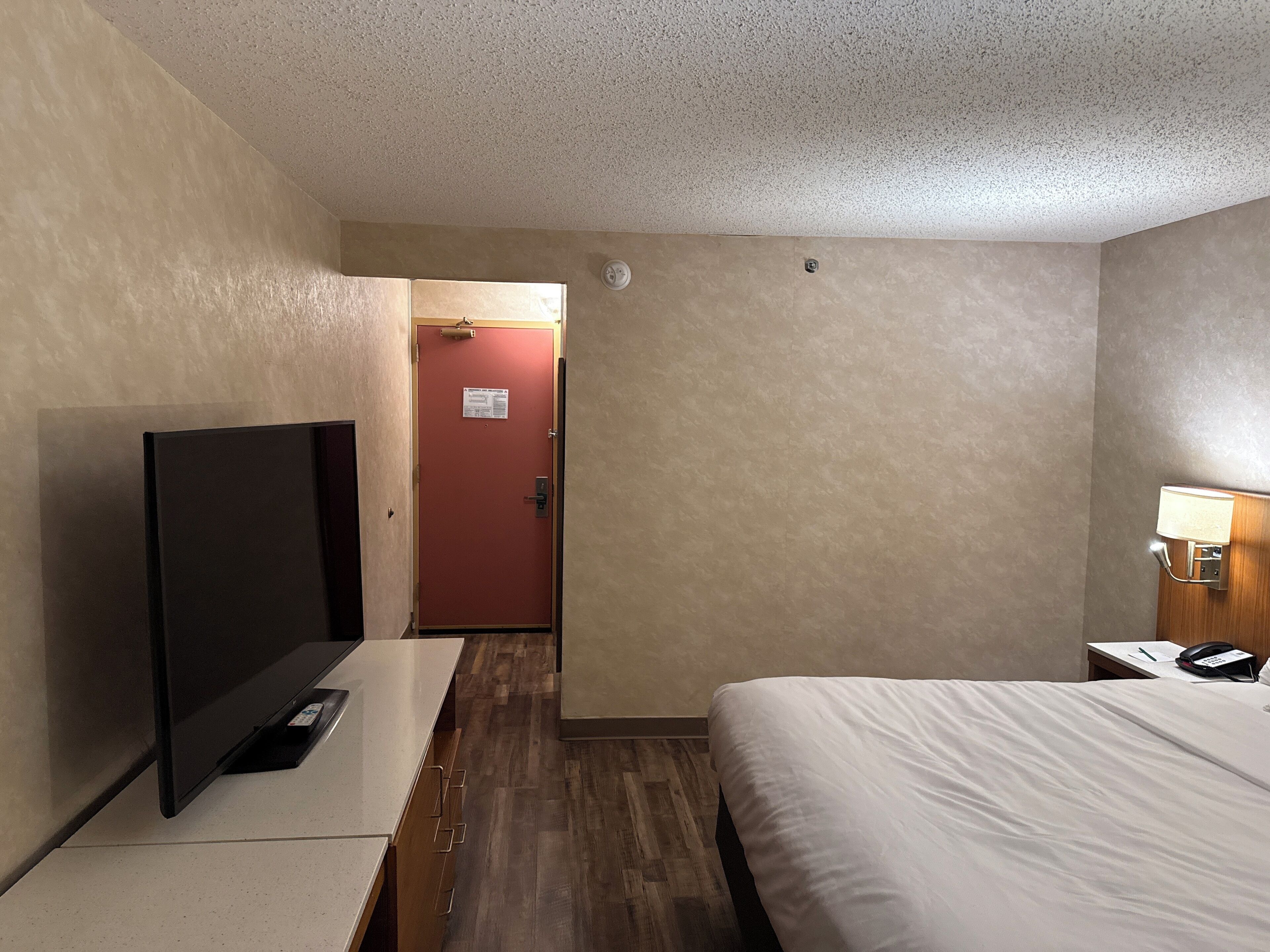 Ett hotellrum med en säng, en tv, ett skrivbord med en lampa och en dörr som leder till ett annat rum.